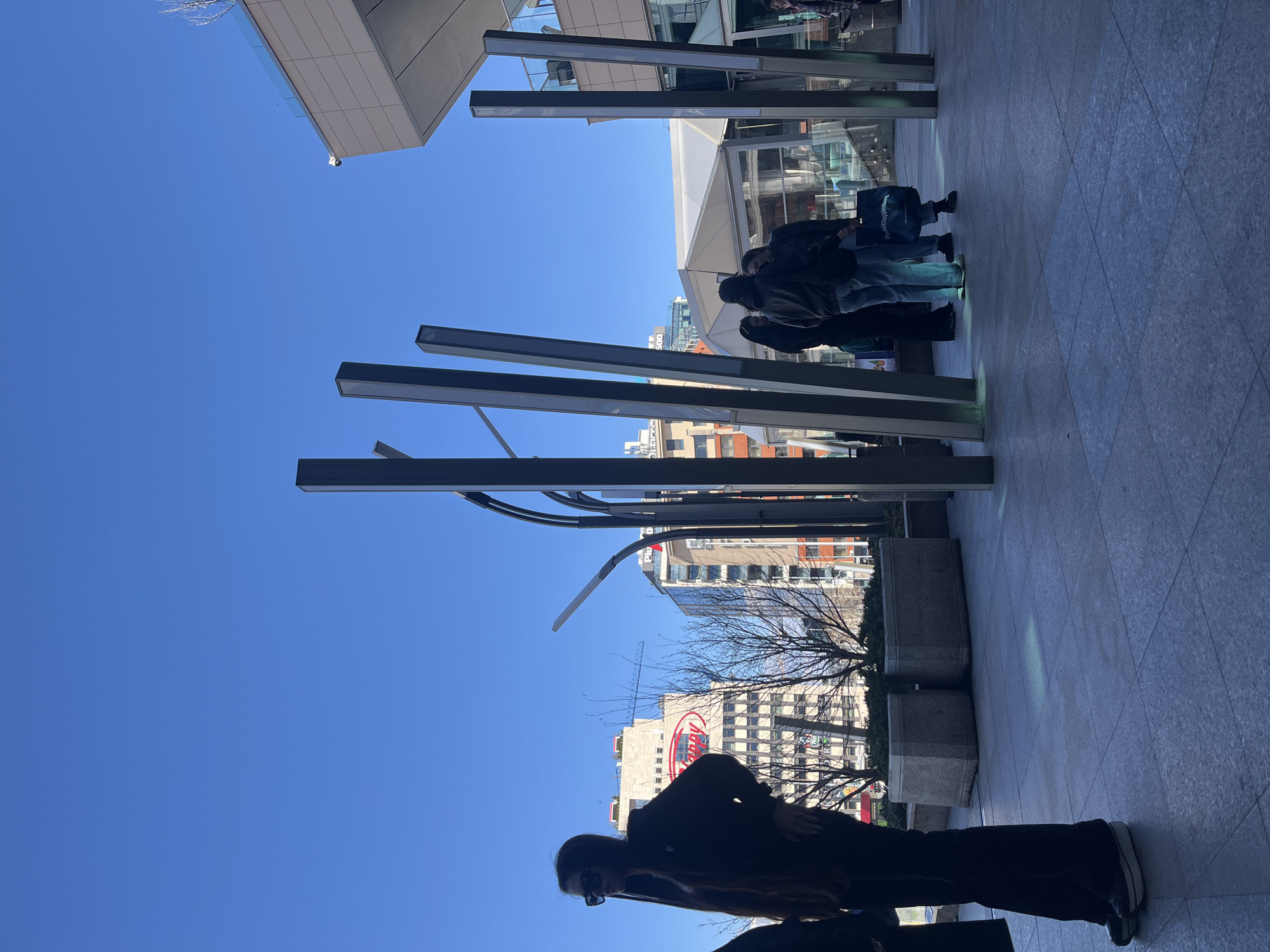 The photo captures a street-level view in Sofia, Bulgaria, characterized by modern architectural elements. Several people are walking with luggage outside of a modern building with a clear blue sky above. A prominent red and white sign of "Marinella" is visible in the background, which gives a clue about the location, which is most likely near Hotel Marinella. The scene is outdoors with buildings and public art visible.
