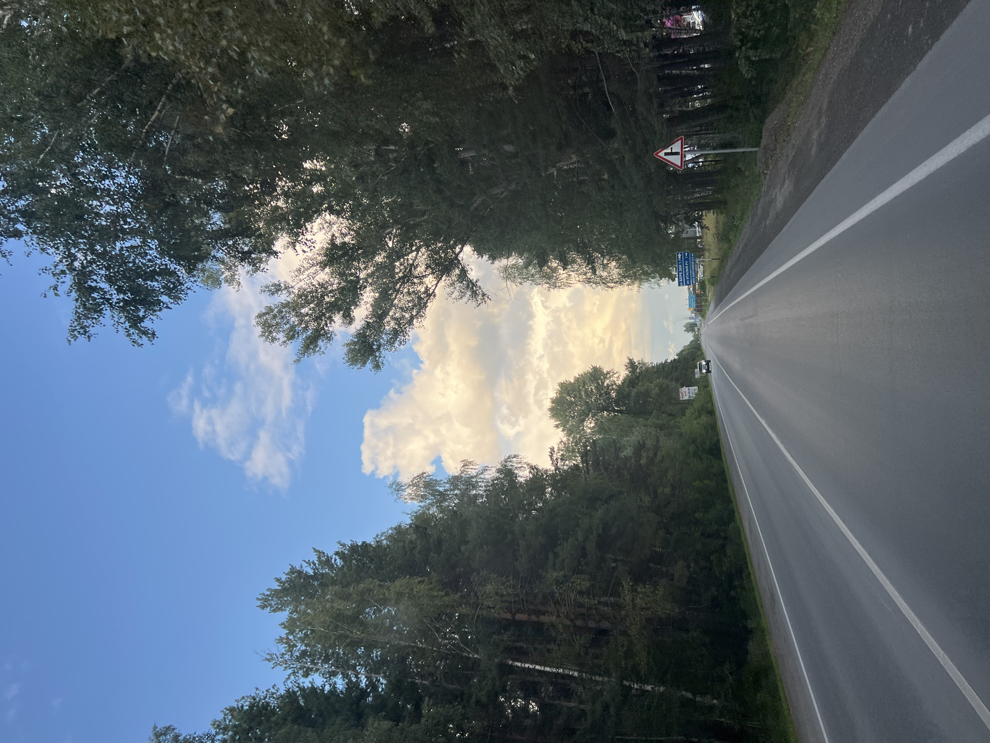 The image shows a paved road with two lanes, bordered by dense trees on both sides. The road is empty except for a distant white vehicle. A traffic sign indicating a junction with a side road is visible on the right side of the road. Several blue signs with white text are also present on the left side, indicating directions to various locations such as "ЮЖНЫЙ" (Southern), "ИТАЛЬЯНСКИЙ РУ" (Italian Ru), and "ВИСК ХИРОШЕВ" (Visk Khiroshev), with distances ranging from 0.5 to 1.7 kilometers. The sky is partly cloudy with the sun setting in the background, casting a warm glow on the clouds. The scene appears to be on the outskirts of Ozernyi, Russia, given the presence of Russian road signs. The overall environment is natural and rural, with a focus on the road and its surrounding forest.