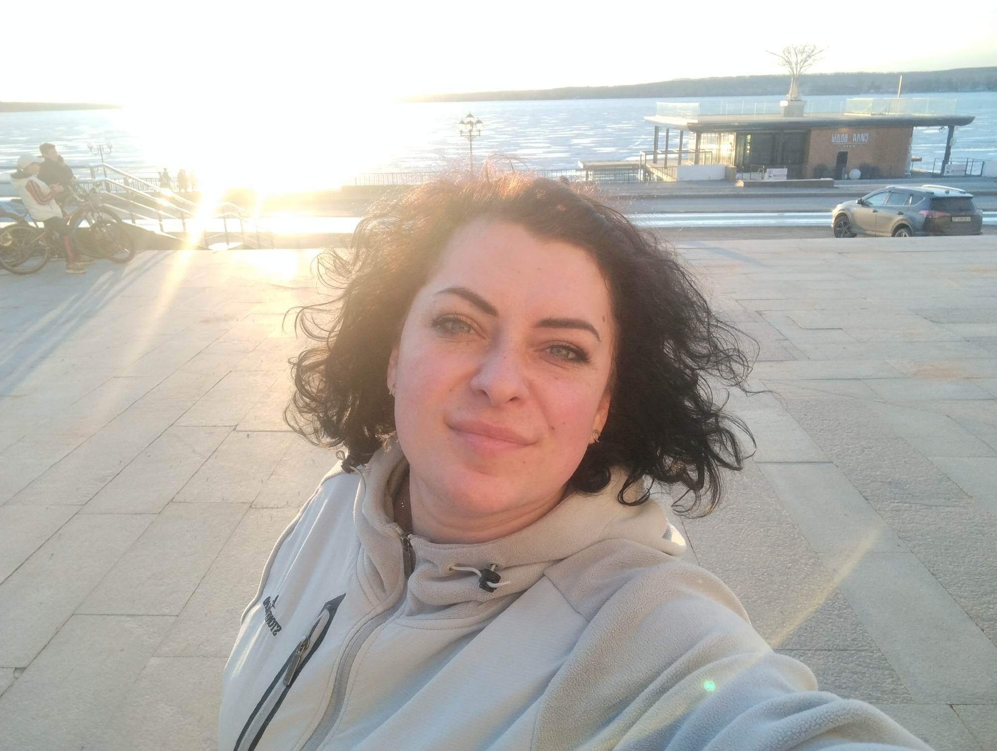A medium shot depicts a woman in a light-colored zip-up fleece jacket. She is looking at the camera with a slight smile. In the background, there is a waterfront scene with a building on the right, a car parked nearby, and people with bicycles to the left. The sun is shining brightly, creating a glare on the water. The setting is a paved area with large stones. Based on the architecture and the water view, the location is Votkinsk, Russia.