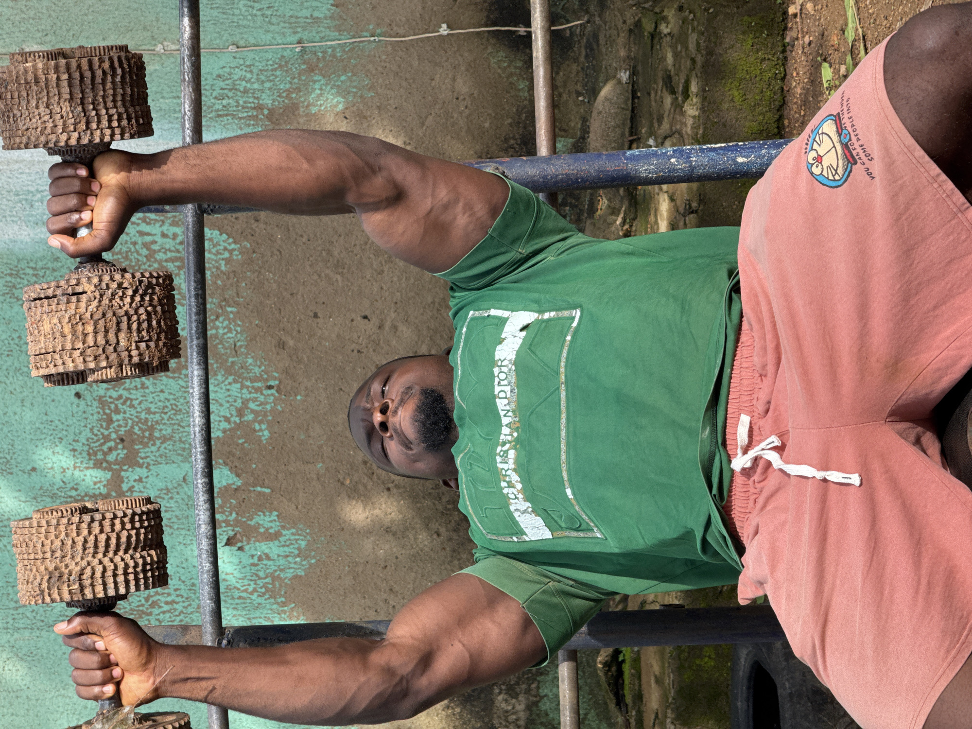 A Black man is pictured in an outdoor setting, performing a chest press exercise with two large, rustic dumbbells. The dumbbells appear to be homemade, constructed from what looks like carved wood or concrete, with a textured surface. The man is seated on an unseen surface, with his arms extended upwards, holding the weights. He is wearing a green t-shirt with a white graphic design and salmon-colored shorts. The background consists of a weathered, green-painted wall and a metal bar, suggesting an improvised gym setup. The ground is dirt and scattered debris. The lighting indicates it is daytime, possibly outdoors in a somewhat dilapidated area, which could be consistent with a less developed part of Akwanga, Nigeria, where access to professional gym equipment might be limited. The man’s muscular physique is evident, particularly his arms and chest.