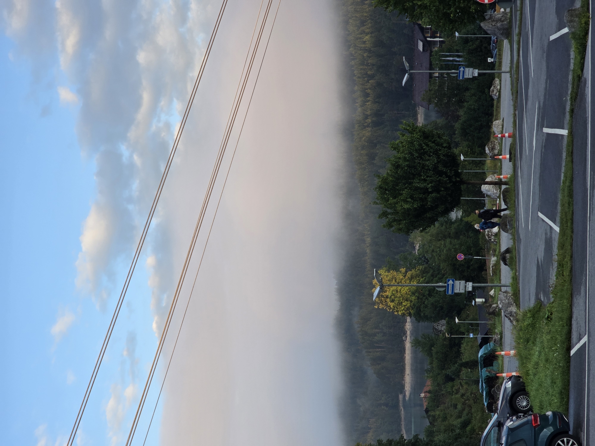 The image depicts a parking lot area at the foot of a mountain range, partially obscured by fog, in Grainau, Germany. In the foreground, several parked cars are visible, including a dark grey sedan and a dark green SUV. The parking lot itself is paved with asphalt and marked with white lines. To the left of the parking lot is a grassy area with some rocks.
In the middle ground, there are several streetlights, some with solar panels. A few traffic cones are placed on the ground. Two individuals are walking away from the camera towards the right side of the image. Behind them, a dense forest covers the lower slopes of the mountains.
The background is dominated by the mountain range, with a thick layer of fog obscuring the upper portions and the peaks. The sky above is blue with scattered white clouds. The overall impression is that of a cool, misty morning in a natural, mountainous setting.