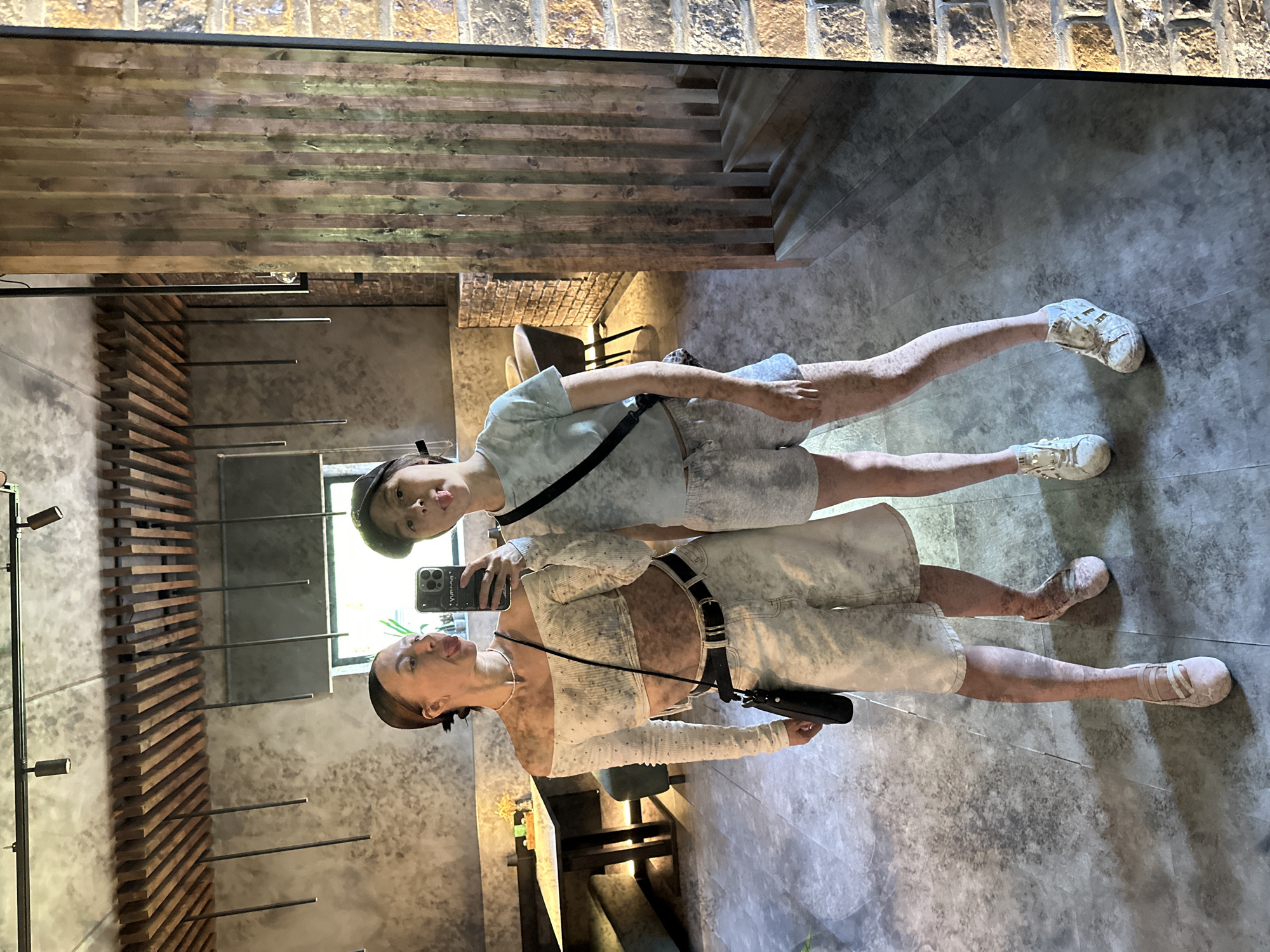 The image shows two people taking a selfie in front of a mirror. The woman is wearing a white outfit with a black belt and is holding a black smartphone. The other person is wearing a t-shirt and a hat. The reflection shows a room with a unique design, including a brick wall, wooden beams, and decorative metal elements. The floor is made of grey tiles. The location is in Votkinsk, Russia.