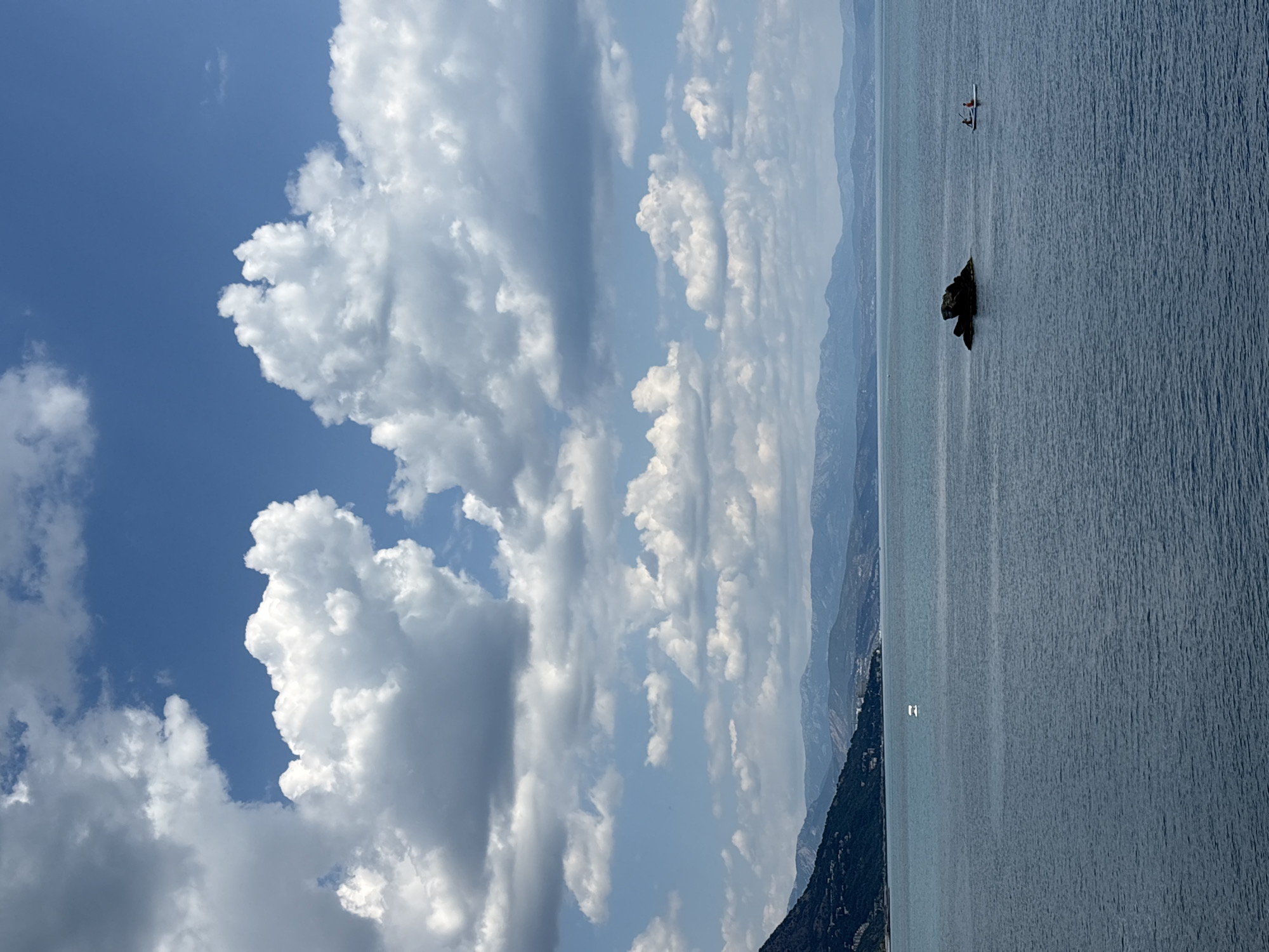 A wide-angle, eye-level shot captures a serene coastal landscape. The foreground is dominated by a vast expanse of calm, deep blue water, its surface slightly rippled. In the middle ground, a rocky outcrop emerges from the water, a dark, rugged shape against the lighter sea. To the right of this rock, two individuals are paddling on a single white kayak, their figures small against the immensity of the water.
Further in the distance, a white buoy with a visible structure is anchored in the sea. The background features a sloping shoreline with dark green, densely vegetated hills dotted with buildings. Beyond the hills, a range of hazy, layered mountains stretches across the horizon, their peaks softened by distance and atmospheric conditions.
The sky above is a vibrant blue, dramatically filled with a mix of large, fluffy white cumulus clouds and smaller, scattered white clouds. Some clouds are tinged with grey, suggesting their depth and density. The lighting indicates a bright, overcast day, with diffused sunlight creating soft shadows and a consistent illumination across the scene. The overall impression is one of tranquility and natural beauty.
This image does not depict Kiparisnoe, Ukraine.