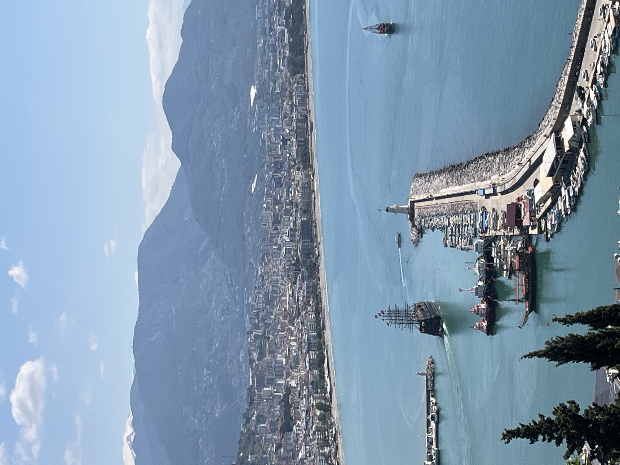 This high-angle, panoramic image captures a vibrant coastal scene, characteristic of Alanya, Turkey. On the left, towering, dark mountains dominate the background, their rugged slopes meeting a sprawling urban landscape. Numerous buildings, mostly light-colored, extend along a narrow, sandy beach that borders the clear, light blue sea.
To the right, a busy harbor is the focal point. A long, curved stone breakwater, providing protection for the port, is densely populated with a variety of boats and yachts moored along its inner edge. A prominent white lighthouse stands at a bend in the breakwater, overseeing the maritime activity. Within the calm harbor waters and just beyond, several vessels are visible. A large, multi-masted replica pirate ship, dark-hulled and adorned with red flags, is a striking feature, leaving a subtle wake as it maneuvers. Smaller speedboats and yachts also dot the water, some docked and others moving.
The sky overhead is a bright, clear blue with scattered white clouds, indicating a sunny day. The natural light casts soft shadows, suggesting it is late morning or early afternoon. The scene conveys a sense of bustling activity within a picturesque Mediterranean setting, combining the natural grandeur of mountains and sea with the dynamic life of a port city. There is no visible text in the image.