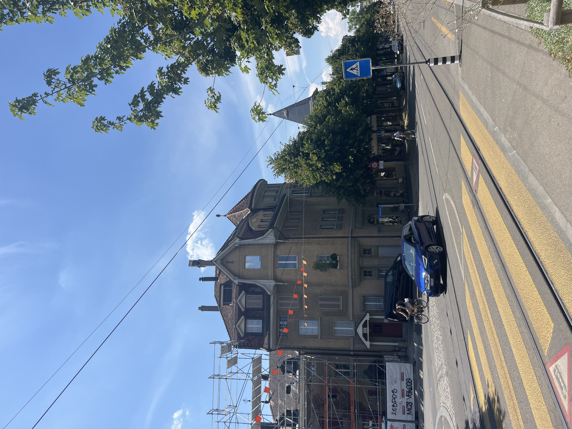 This image captures a street scene in Bern, Switzerland, on a sunny day. In the foreground, a wide pedestrian crossing with yellow stripes is visible, with tram tracks running parallel to it. A blue sedan is driving across the intersection, and a cyclist is riding past on the right side of the road.
The buildings lining the street are multi-story structures with traditional European architecture. The building on the left is partially covered in scaffolding, indicating ongoing construction or renovation. Orange triangular flags are strung across the facades of some buildings. Trees with lush green foliage line the sidewalks, providing shade.
A blue pedestrian crossing sign is prominently displayed on a pole. In the background, a church spire with a dark roof can be seen peeking above the trees. Power lines crisscross the sky, which is mostly clear with a few wispy clouds. The overall impression is a vibrant urban environment with a blend of old and new architectural styles.