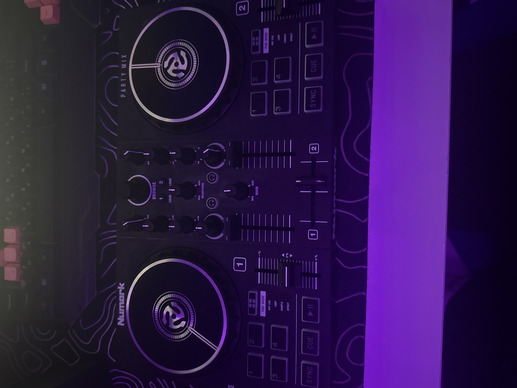 This is a top-down, medium shot of a Numark Party Mix DJ controller. The controller is black with purple topographic-like graphics. The device is illuminated by purple lighting, casting shadows. The DJ controller features two jog wheels, sliders for volume and pitch, various buttons for cueing, looping, and effects, and a central browse knob. Above the DJ controller, a portion of a keyboard with pink keys is visible. The setting is indoors, on a flat surface, likely a desk or table. The scene does not contain any people or specific identifying landmarks of Burgas, Bulgaria.
