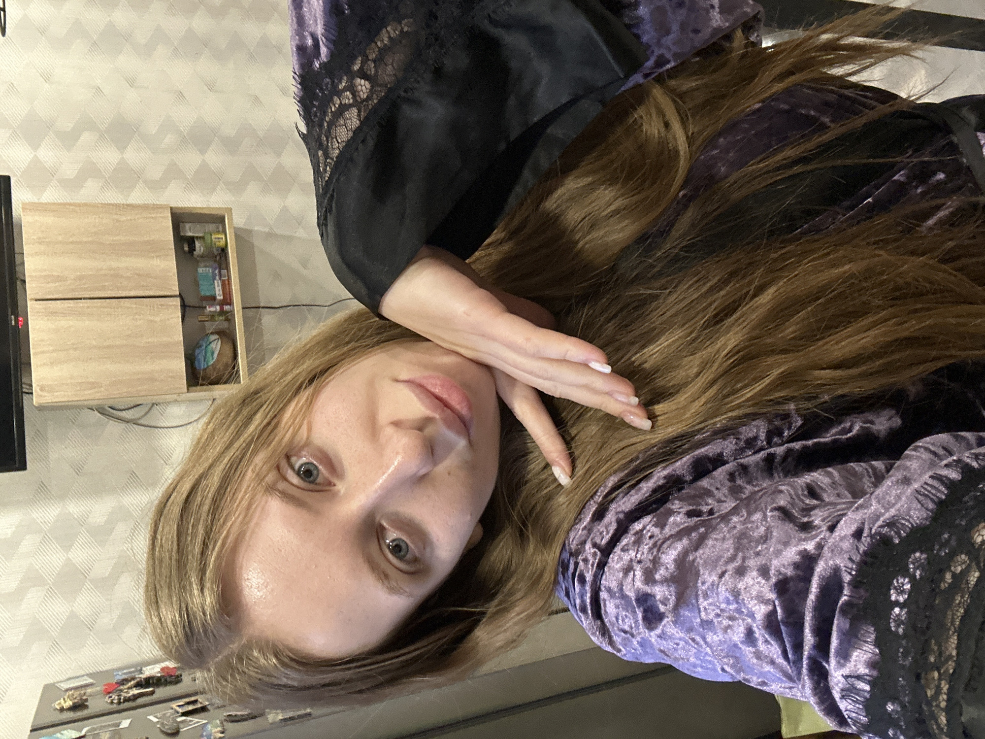 This media file is a portrait of a young woman, captured indoors, and is rotated 90 degrees clockwise. The woman, with fair skin, light brown hair, and blue eyes, looks directly at the camera with a neutral, slightly relaxed expression. Her right hand, adorned with light-colored nail polish, rests gently against her cheek. She is wearing a dark purple velvet robe or dress, detailed with black lace trim on the sleeves and shoulders.
The background suggests a domestic setting. To the left and below her, a grey wall with a subtle textured diamond pattern is visible. Against this wall, a light wooden cabinet or shelf unit features an open compartment holding various small items. Partially visible on the far left is a black electronic device, possibly a television, displaying the red digits "99:". Below the cabinet, a metallic surface, likely the top of a refrigerator, is decorated with several small magnets. The woman is posing for the photograph, and no specific activity is taking place beyond this.