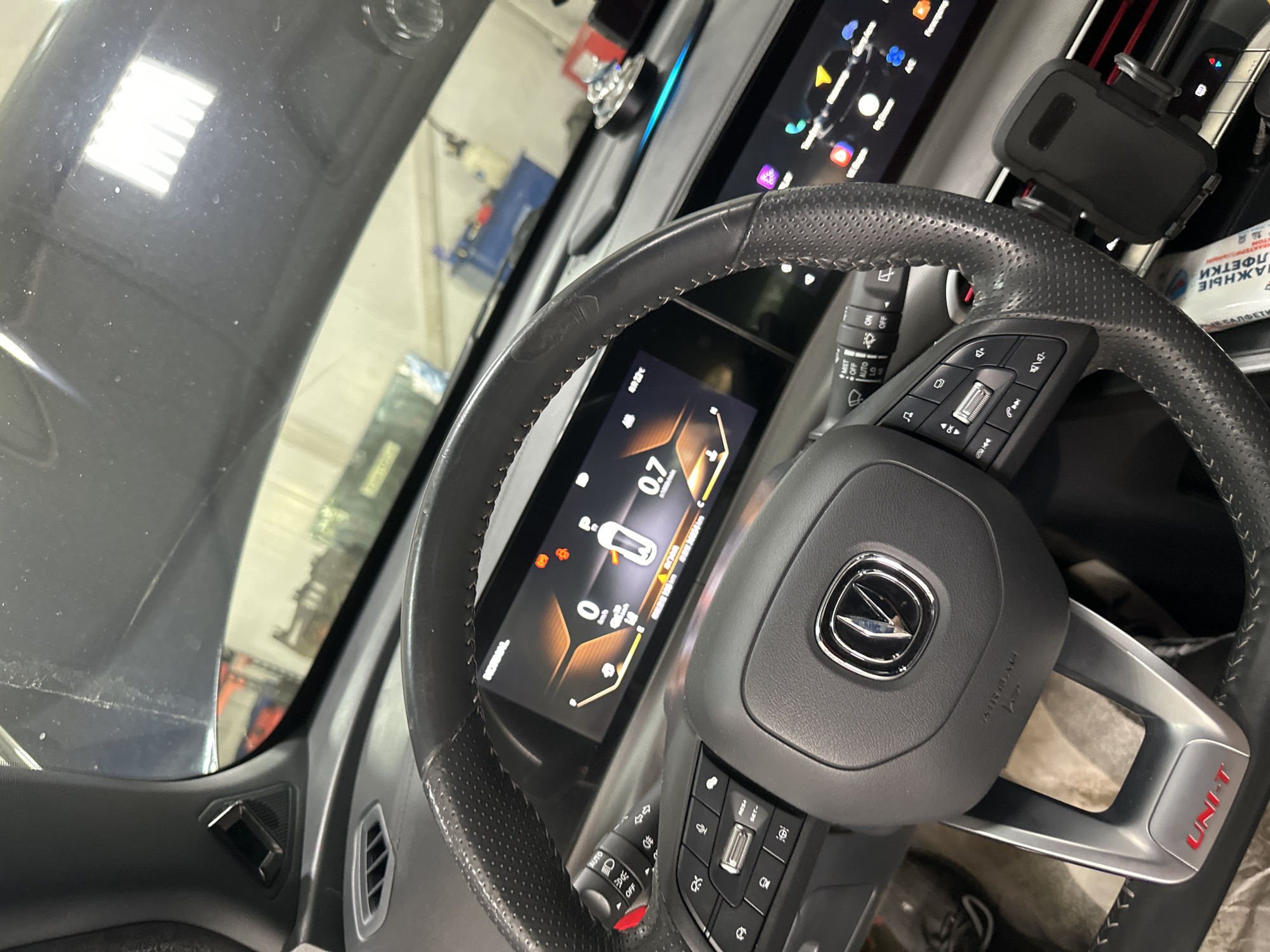 The image shows the interior of a car, likely a modern vehicle with a digital dashboard and steering wheel controls. The dashboard displays various icons and information, including what appears to be speed, battery level, and tire pressure. The steering wheel is leather-wrapped and has buttons for media control and cruise control. A mobile phone holder is attached to the right side of the steering column. The overall environment appears to be a workshop or garage, with equipment and machinery visible in the background through the windshield. The image is a close-up shot focusing on the driver's area of the car. The presence of dust on the dashboard and a slightly cluttered environment suggests the car may be undergoing maintenance or is in a less-than-pristine state. No people are visible in the image. It is impossible to determine the exact location within Blagoveshchensk, Russia based on the visual information provided.