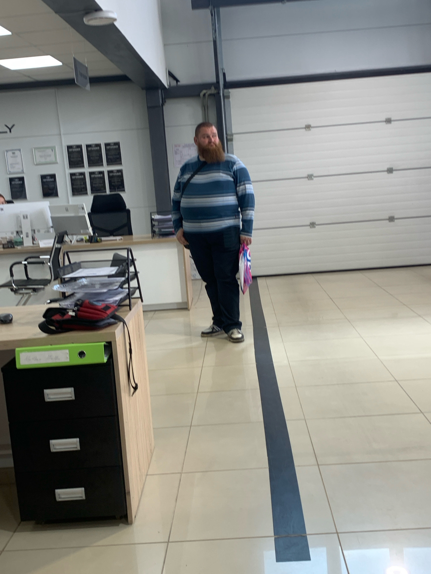 In the city of Чыжэвіцкі сельскі Савет, Belarus, a full-bodied man with a reddish-brown beard stands in what appears to be an office or commercial space. He is wearing a horizontally striped sweater in shades of blue and a dark-colored pair of pants. The man is holding a multi-colored plastic bag in his right hand and has a dark strap across his shoulder. He is positioned in front of a large, white, sectional garage door. To his left, there is a desk with office supplies, including papers, a computer monitor, and a black office chair. A three-drawer black filing cabinet with silver handles is visible in the foreground to the left, topped with a green binder and a black object with a red accent. The floor is tiled with light-colored square tiles and features a dark grey line running vertically in the center of the image, separating the space. The environment suggests an indoor setting with bright overhead lighting.