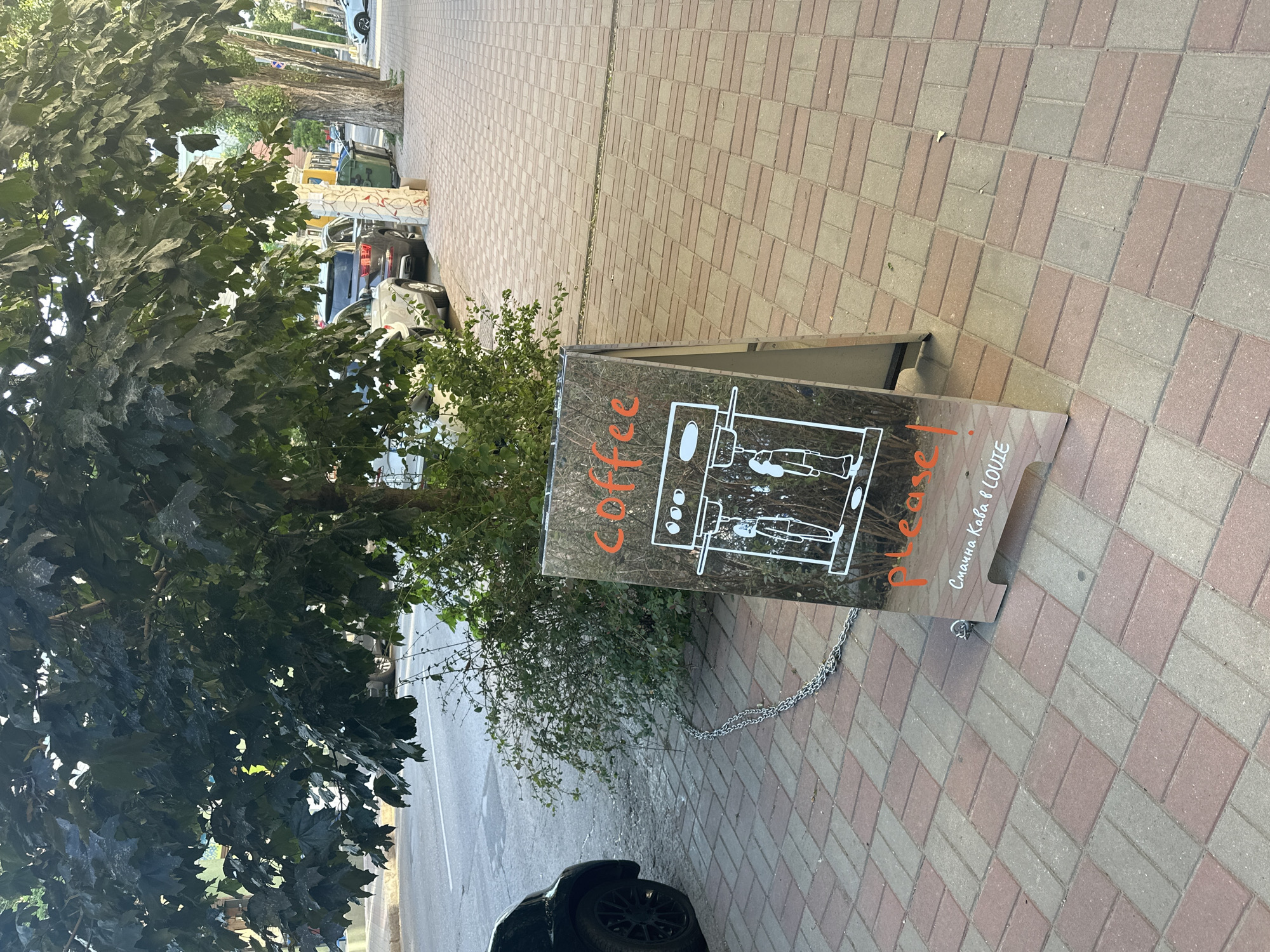 A sidewalk sign for a coffee shop stands on a paved walkway with red and tan brickwork. The sign is a dark, reflective material with an A-frame structure and is anchored by a silver chain.
On the sign, the word "Coffee" is written in orange cursive script. To the right of "Coffee," a simple white line drawing depicts an espresso machine. Below the machine, there are two figures, one of whom appears to be handing a cup to the other. Below this drawing, the word "Please!" is written in orange cursive, mirroring the style of "Coffee." Further down, in smaller white cursive, the text reads "Смачна Кава в LOUVE," which translates to "Delicious Coffee at LOUVE."
The sign is positioned next to a large, leafy green tree and some smaller bushes. In the background, cars are parked along the street, and a road is visible. The scene appears to be outdoors during daylight. The overall impression is one of a casual, inviting coffee shop advertisement on a pleasant day.