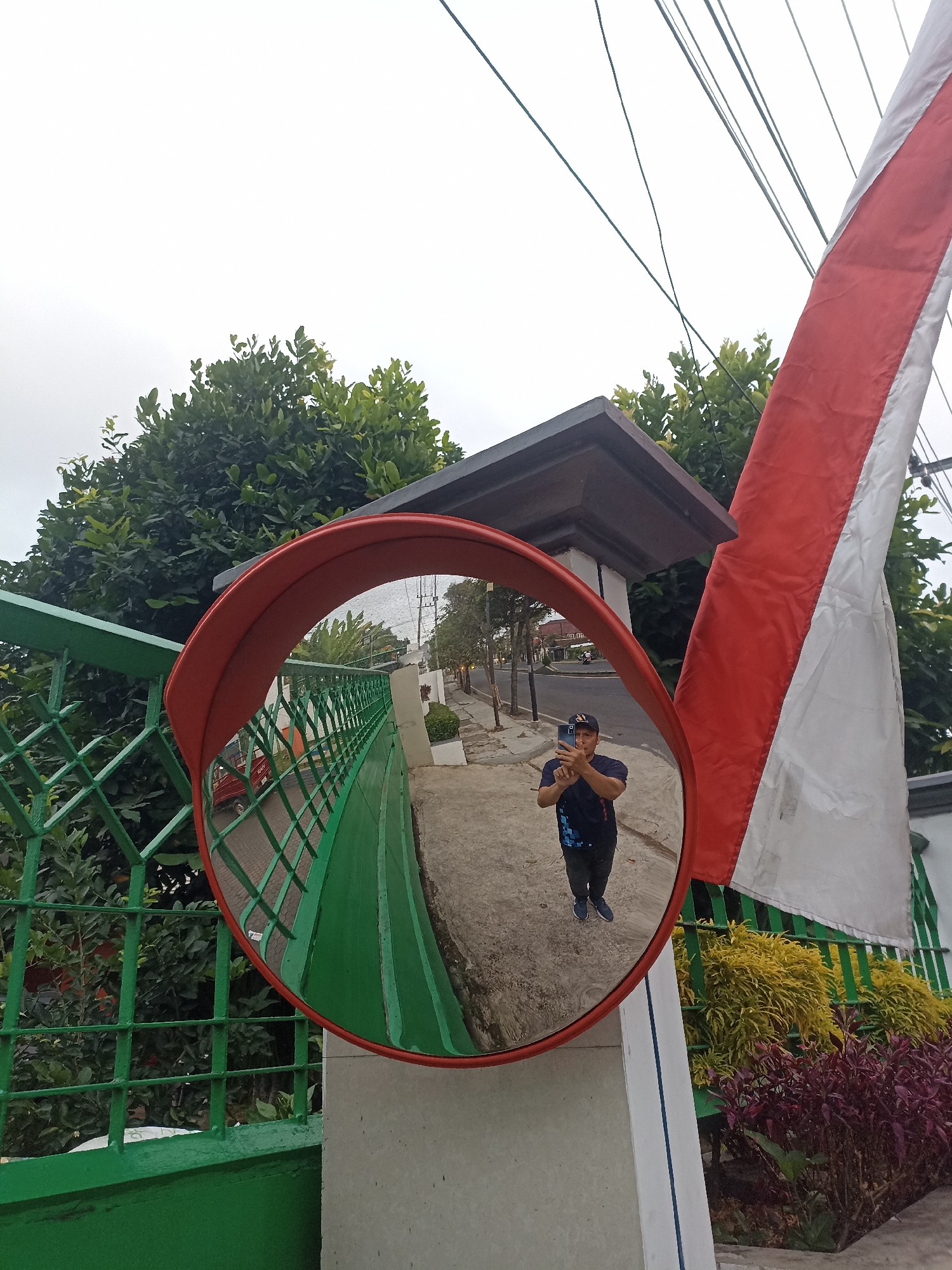 A man takes a selfie in a convex mirror mounted on a white concrete pillar next to a green metal fence. The mirror reflects the man, who is wearing a dark blue t-shirt with a blue geometric pattern, a dark baseball cap, and dark pants, holding a smartphone to take the picture. He is standing on a paved sidewalk. To the left of the mirror, a green fence encloses lush green foliage. Behind the fence, a red vehicle is visible on a street. To the right of the pillar, a red and white flag, likely the Indonesian flag, is draped. Further to the right, there are decorative plants with yellow and purple foliage. The background shows trees, buildings, and utility wires against an overcast sky. The location is on a street in Banyuwangi, Indonesia, as indicated by the Indonesian flag.