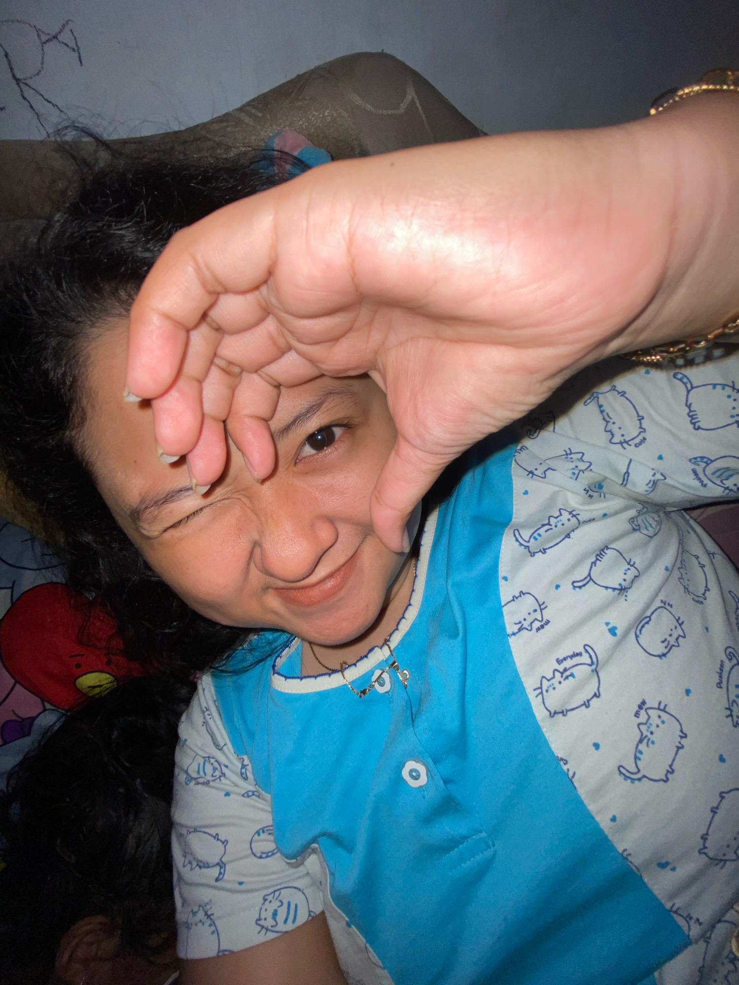 A selfie taken by a person wearing a blue and white pajama top with a cat print. The person is winking and making a heart shape with their fingers over their face. The background appears to be a bed with a pillow. The image is taken from a close-up perspective, showing the person's face and upper body.