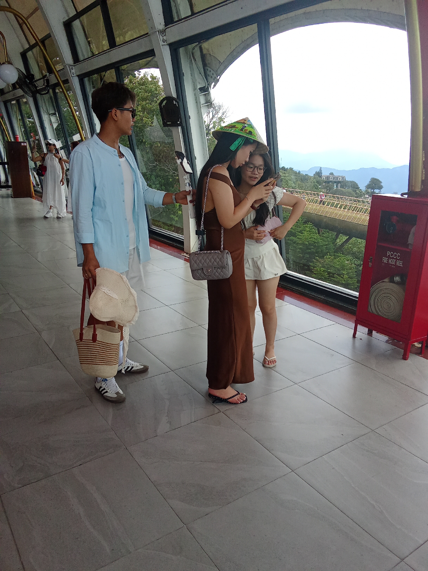 The image depicts three individuals indoors, standing near a large window overlooking a mountainous landscape with structures and greenery. In the foreground, a person with short dark hair and sunglasses wears a light blue open-collared shirt over a white t-shirt and white shorts. They are holding a woven straw tote bag with red straps in their left hand and a straw hat in their right hand. To their right, a person wearing a dark brown dress with a quilted grey handbag on their shoulder is looking down at a smartphone. To the right of them, a person in a white top and white skirt, wearing a conical hat, also looks at the smartphone. The floor is tiled in a light grey stone pattern. In the background, there's a yellow railing, trees, and buildings. A red fire hose reel cabinet is visible on the right side of the frame. This scene is consistent with the observation deck at Ba Na Hills, a popular tourist destination in Đà Nẵng, Vietnam, known for its French colonial architecture and panoramic views.