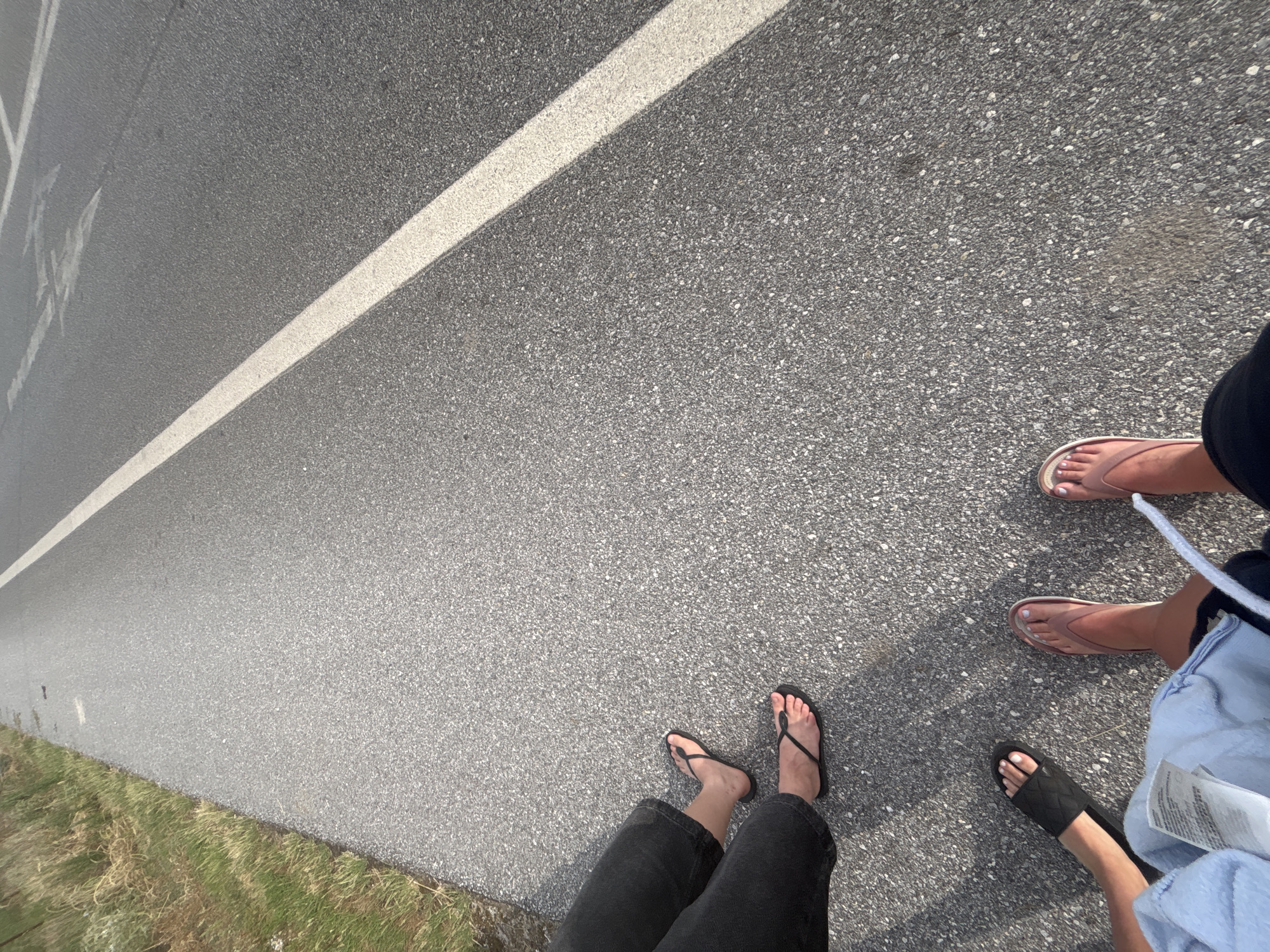 The image shows a low-angle perspective of four pairs of legs and feet on an asphalt road in Prosotsani Municipal Unit, Greece. The road has a white line marking its edge and appears to be a thoroughfare. To the left of the road is a grassy verge with dry vegetation. The individuals are wearing casual footwear, including flip-flops and slides. One individual is wearing dark jeans, another is partially visible with a light blue garment. The lighting suggests it is daytime, possibly late afternoon due to the shadows. The scene captures a moment of stillness, with the focus on the feet and the immediate surroundings.