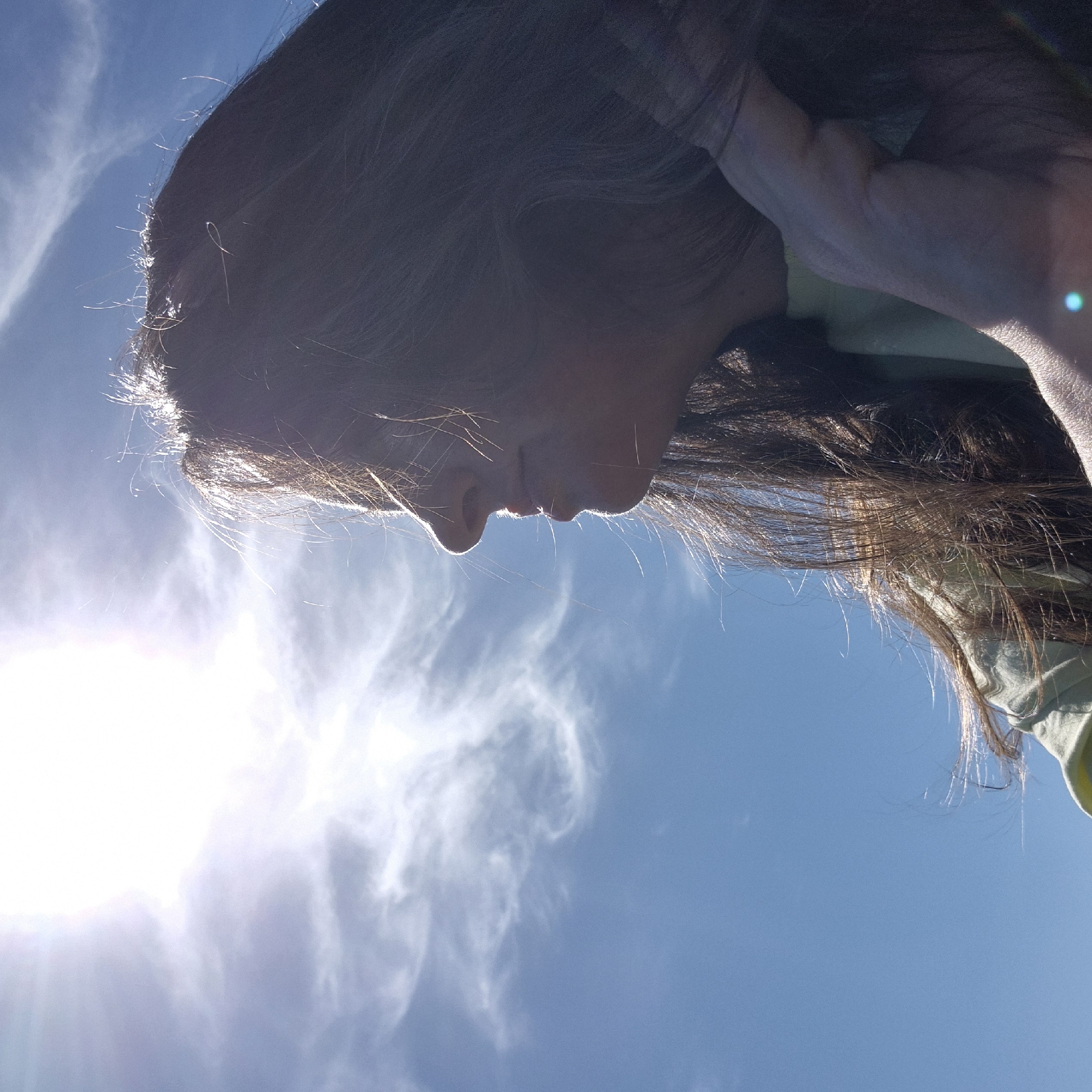 The image is a medium shot, taken from a low angle, of a young woman outdoors against a bright blue sky with wispy clouds and the sun shining intensely. The woman is facing to the left, her profile visible. She has long, dark brown hair that is blowing in the wind and partially obscuring her face. Her skin is illuminated by the bright sunlight, creating a silhouette effect. She is wearing a light-colored garment, possibly a t-shirt. Her right hand is visible near her face, with her fingers gently touching her hair or cheek. The overall impression is one of a serene and windy day. The location is stated as Новостройка, Russia, though there are no specific visual cues in the image to confirm this.