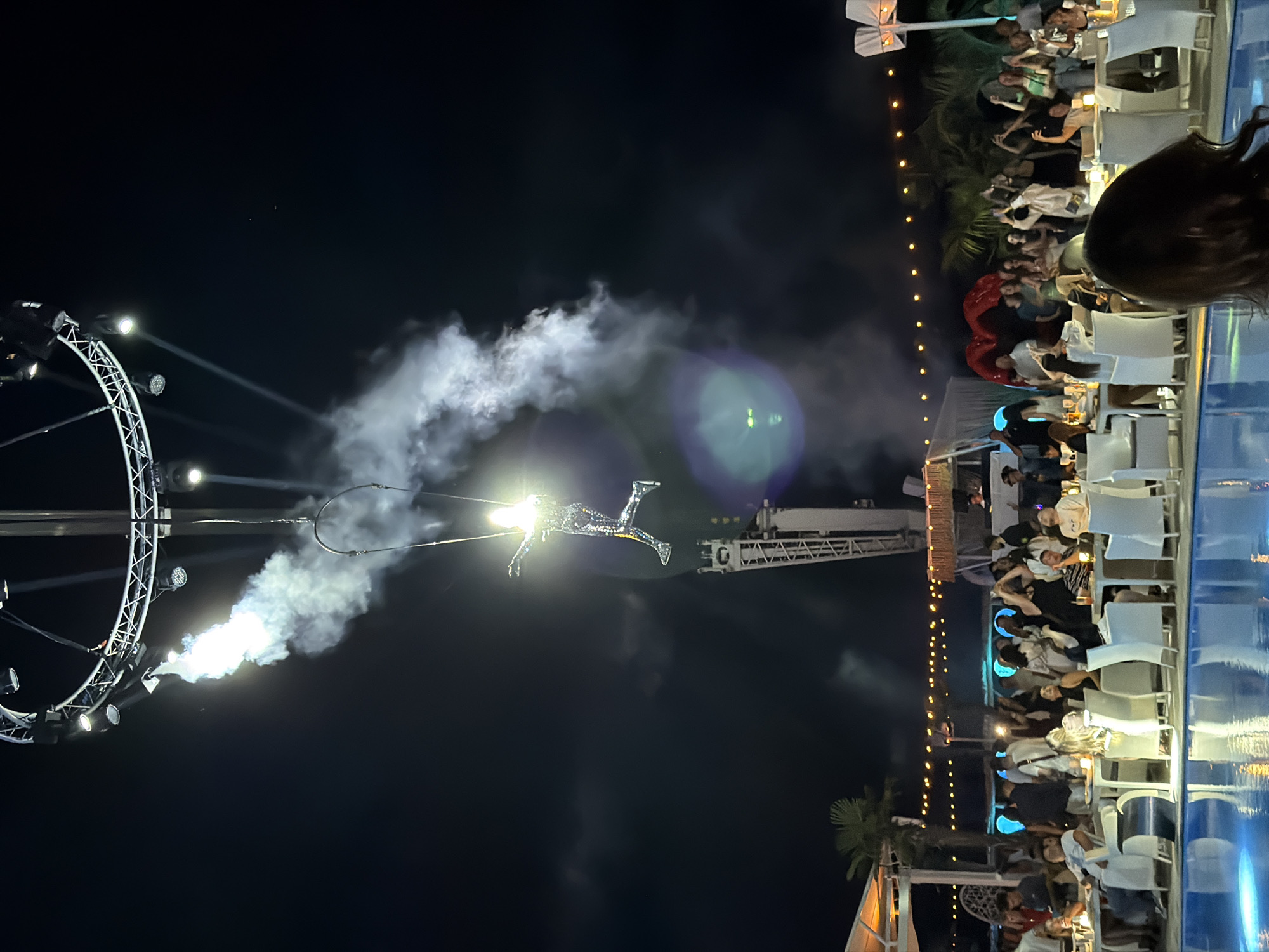 The image depicts an aerial performer in a shimmering costume suspended from a circular rig with spotlights at an outdoor night event. Smoke or mist emanates from the rig, creating a dramatic effect. Below, a crowd of people are seated at tables around a pool, with some standing and dancing in the background. String lights are visible in the distance, illuminating the scene. The setting appears to be a resort or club, possibly near the sea, given the presence of palm trees and the general ambiance. The event seems to be taking place in Odesa, Ukraine, as indicated by the provided context.