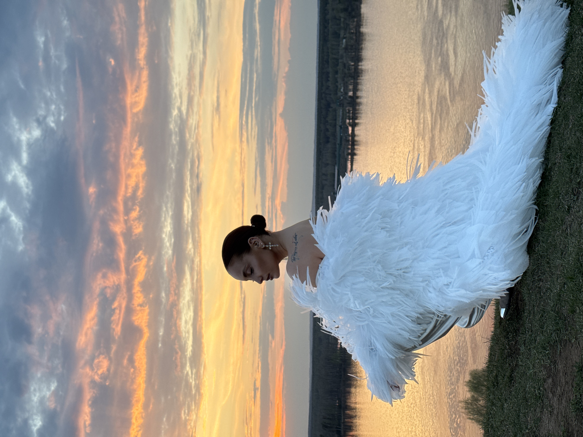 A vertical outdoor photograph depicts a woman in a white, feathery garment sitting near a body of water, likely the location in the city of Ольховка, Russia. She has dark hair pulled back in a bun, wears cross earrings, and has a tattoo on her back. The woman is looking down, and her face is in profile. The backdrop features a sunset with shades of orange and grey clouds. The grass is green near the woman's feet. The water is a shimmering golden color, reflecting the sunset. Trees are visible in the background across the water.