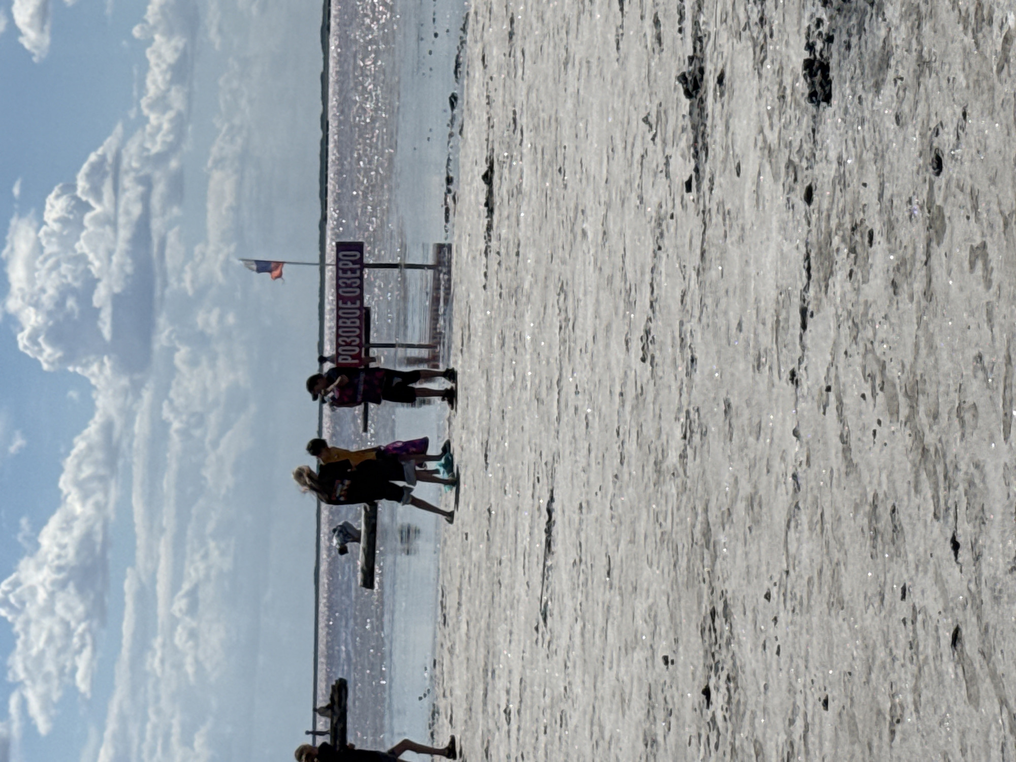 The image shows a sunny day at a salt lake. In the foreground, there is a cracked, dry salt bed with some dark rocks. In the middle ground, there is a pinkish, shallow body of water reflecting the sunlight. Several people are standing near the water's edge. One person is holding a camera. A sign with Russian text that appears to say "РОЗОВОЕ ОЗЕРО" (Pink Lake) is visible. A Russian flag is also flying on a pole behind the sign. The background shows a clear blue sky with scattered white clouds. The overall scene suggests a tourist location at a salt lake.