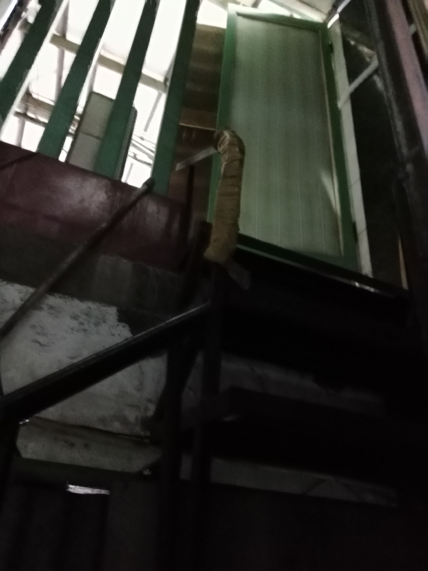 The image depicts a narrow, dimly lit indoor staircase constructed with dark metal railings and a reddish-brown wooden handrail. The stairs themselves are not clearly visible due to darkness. To the left of the staircase, there are vertical green beams and a section of what appears to be a wall or partition made of concrete or plaster, showing some wear and tear. Above the staircase, there are structural elements and a glimpse of a light-colored ceiling or upper floor. To the right, a light green door with vertical paneling is partially visible, slightly ajar. The overall impression is of a functional, perhaps somewhat neglected, staircase in an urban setting. The scene is consistent with the back or service areas of buildings commonly found in areas like West Jakarta, Indonesia, where utilitarian architecture is prevalent. There are no people or clear actions discernible in the image.