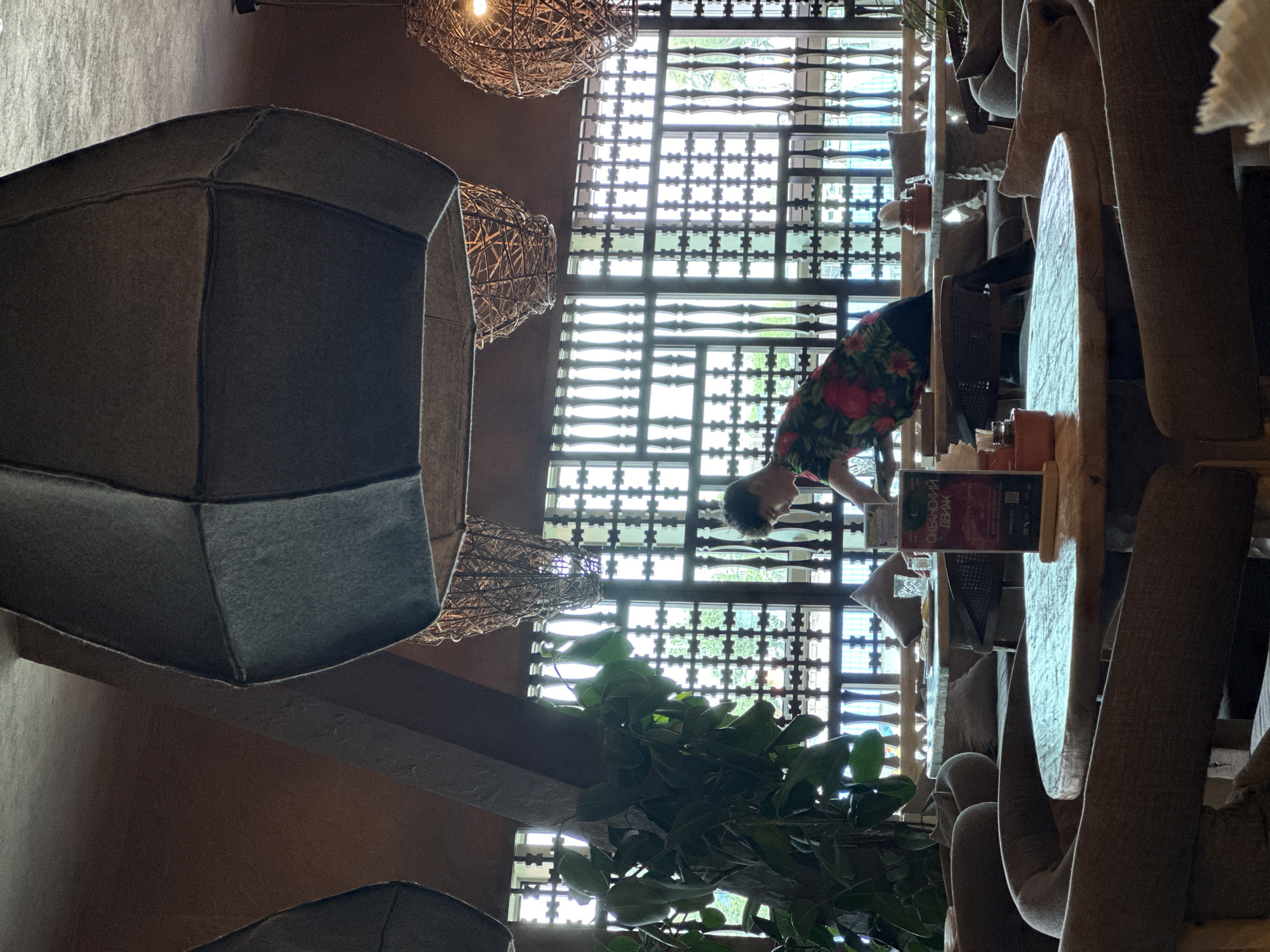 A person wearing a floral shirt is seen reaching for something on a table in a restaurant. The restaurant interior features large, decorative hanging lamps, wooden lattice window coverings, and comfortable seating arranged around wooden tables. A large potted plant is visible in the foreground. The overall ambiance is dimly lit, with natural light filtering through the windows.