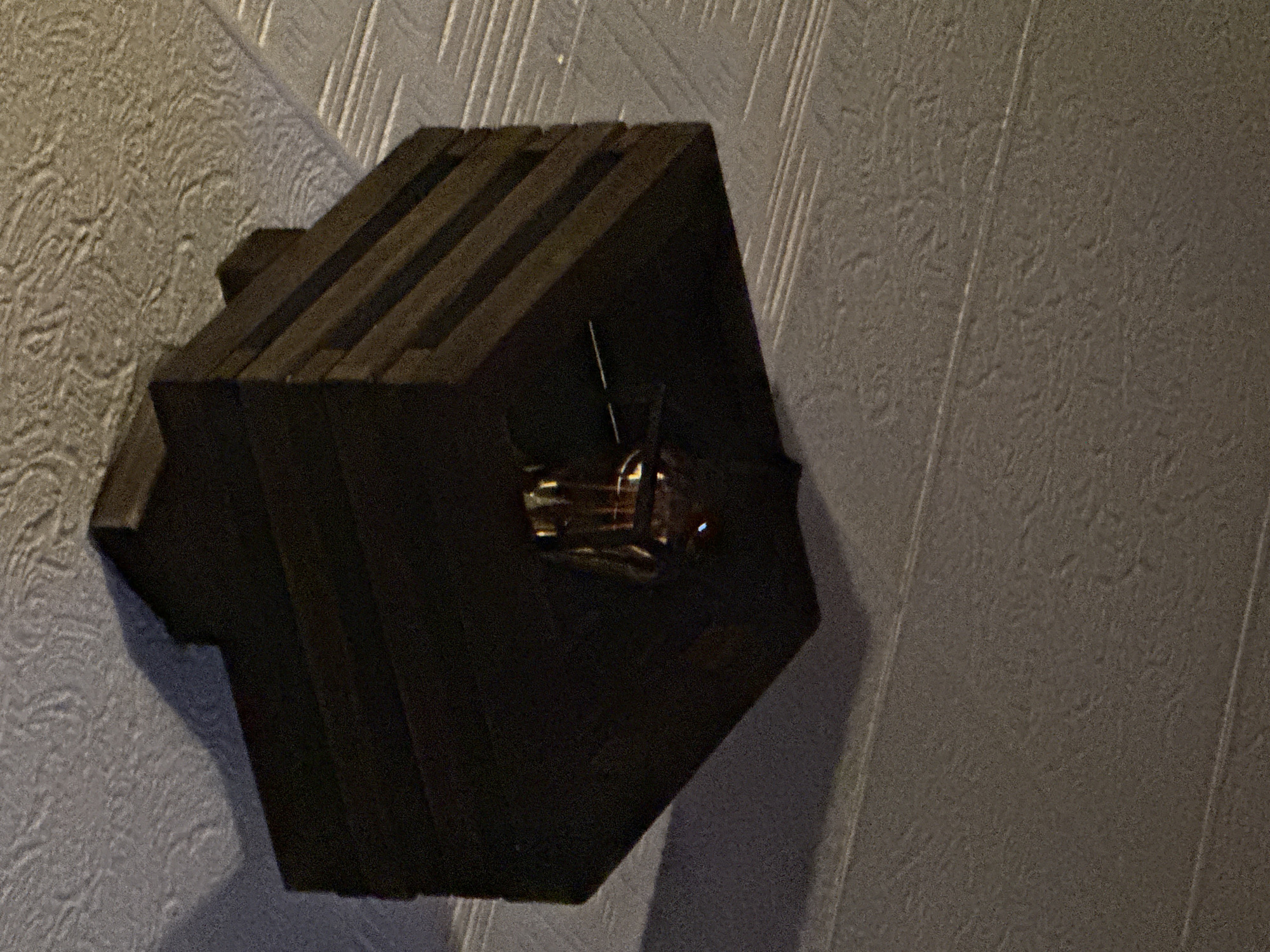 This image shows a close-up, low-angle view of a wooden light fixture mounted on a textured ceiling. The fixture is dark brown, appears to be made of several horizontal planks, and has a tiered, somewhat boxy shape. Suspended from the center of the fixture is a glass light bulb, which is illuminated and casts a warm glow. The ceiling is a light color with a raised, patterned texture, possibly popcorn or stucco. The overall lighting suggests an indoor environment, likely residential or commercial. There is no indication of the city of Galinė, Lithuania, within the image itself.