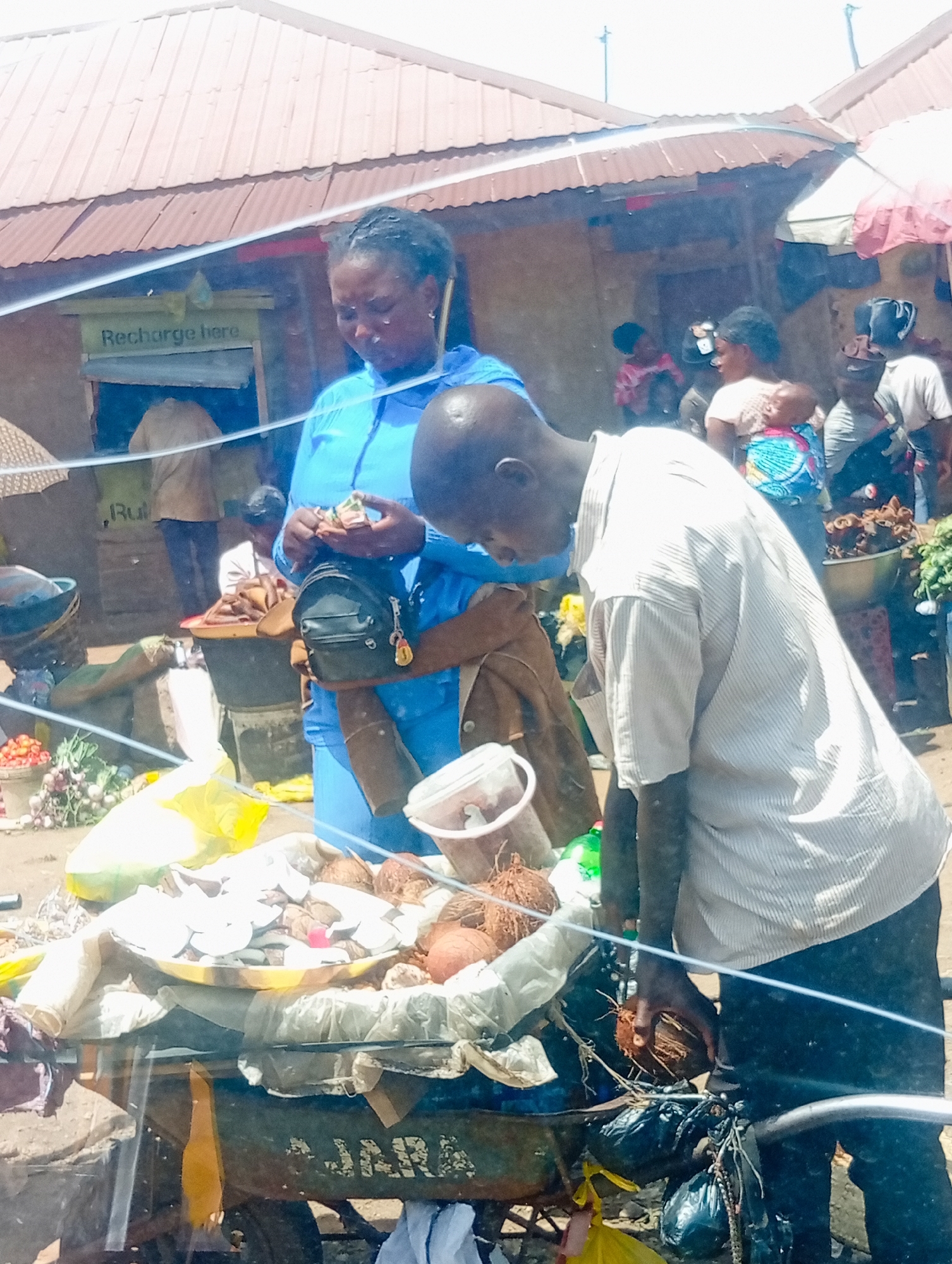 The media file depicts a bustling outdoor market scene in Bukuru, Nigeria. In the foreground, a man wearing a light-colored, pinstriped shirt and dark trousers is leaning over a cart laden with coconuts. He is holding a coconut in his right hand, appearing to inspect or sort it. A woman, dressed in a bright blue top, stands next to him, also interacting with items on the cart. She is holding what appears to be money or small goods in her hands and has a dark shoulder bag.
Behind them, various market stalls are visible, with corrugated metal roofs suggesting a typical informal market structure. The background reveals more people, including children and other vendors, some with goods displayed. The overall atmosphere is one of activity and commerce.
Notable details include a sign in the background that reads "Recharge here," indicating a service available at the market. The lighting suggests it is daytime, with bright sunlight casting shadows, implying fair weather. The presence of fresh produce and coconuts points to a food market. The expressions on the faces of the individuals are difficult to discern clearly due to the image quality, but the scene conveys a sense of everyday life and economic activity. The cart in the foreground has faded white text that appears to read "AJARA".
