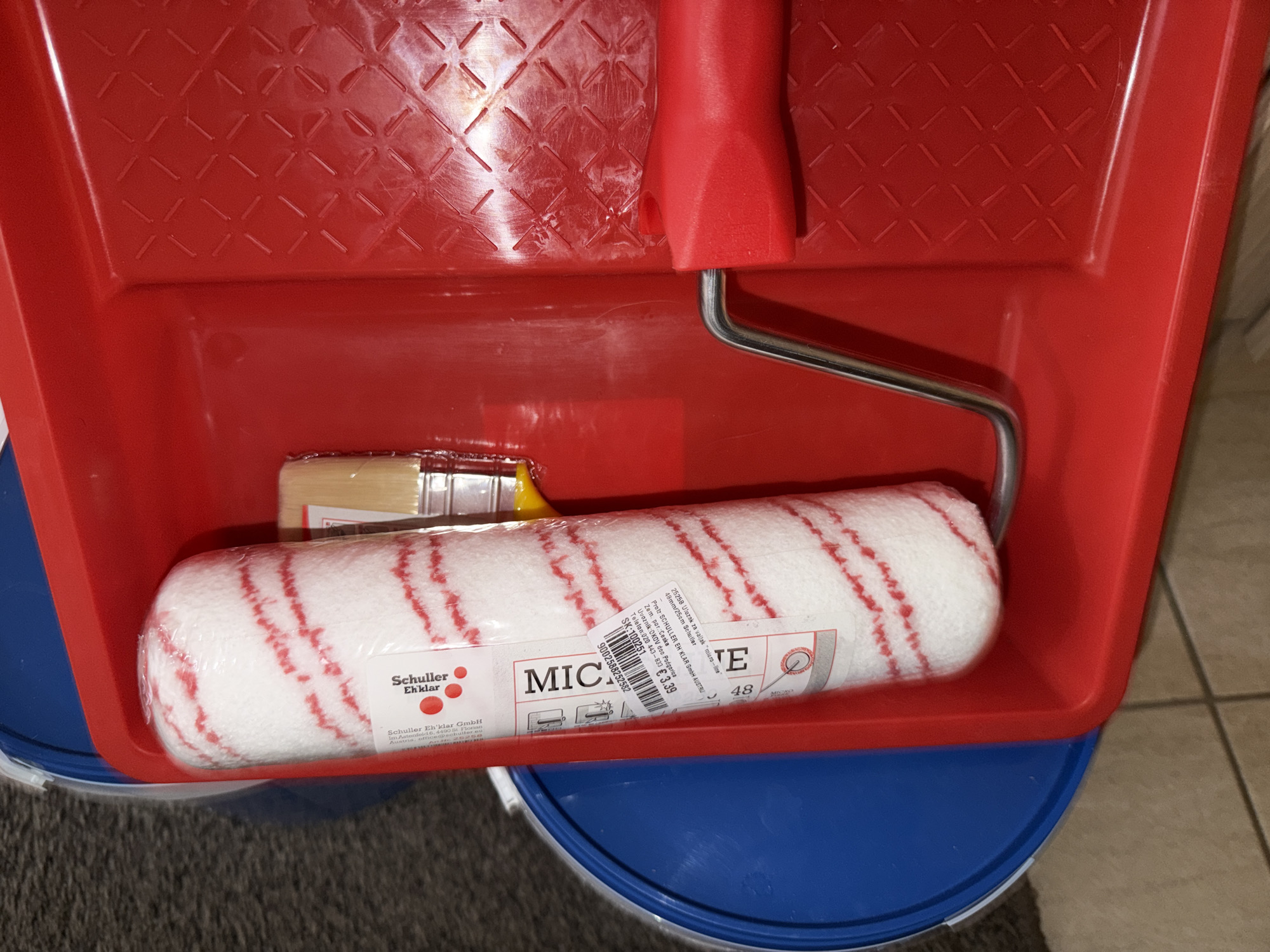 The image shows a red paint roller tray containing a paint roller and a brush. The paint roller is white with red stripes and is still wrapped in plastic. The brush has a wooden handle and light-colored bristles. The tray itself has a textured surface in the bottom right section. In the background, a blue bucket is partially visible. The items appear to be new and unused. The scene is indoors, possibly a home or a storage area. There is no indication of the location in Budva, Montenegro.