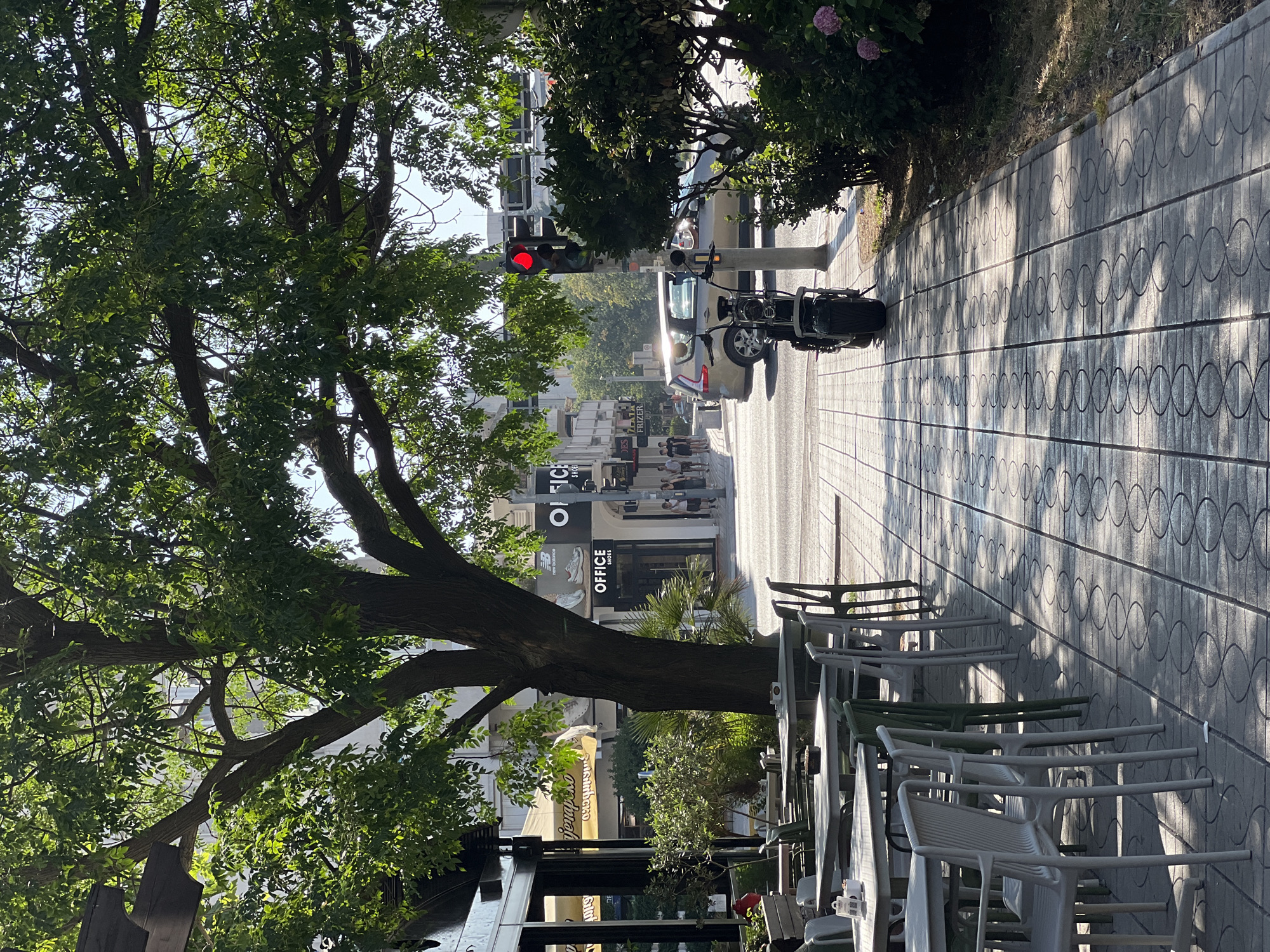 The image captures a street scene in Bar, Montenegro, on a sunny day. The perspective is from an outdoor seating area of a cafe or bar, looking down a street lined with buildings and trees.
In the foreground, to the right, are several white plastic tables and chairs, some stacked. A large tree with lush green foliage dominates the left side of the image, its branches reaching across the frame.
Further down the street, a traffic light is visible, showing a red signal. A silver car is stopped at the light, and behind it, a motorcycle is parked on the sidewalk. The sidewalk itself is paved with patterned tiles that cast shadows from the sunlight.
The buildings lining the street appear to be commercial establishments, with visible signage. One sign reads "OFFICE SHOES". Another sign is partially obscured but appears to be yellow and has text that might say "Jezerska". People are visible walking on the sidewalk in the distance.
The overall impression is a bright, possibly late afternoon scene, given the long shadows cast by the trees and buildings. The weather is clear and sunny.