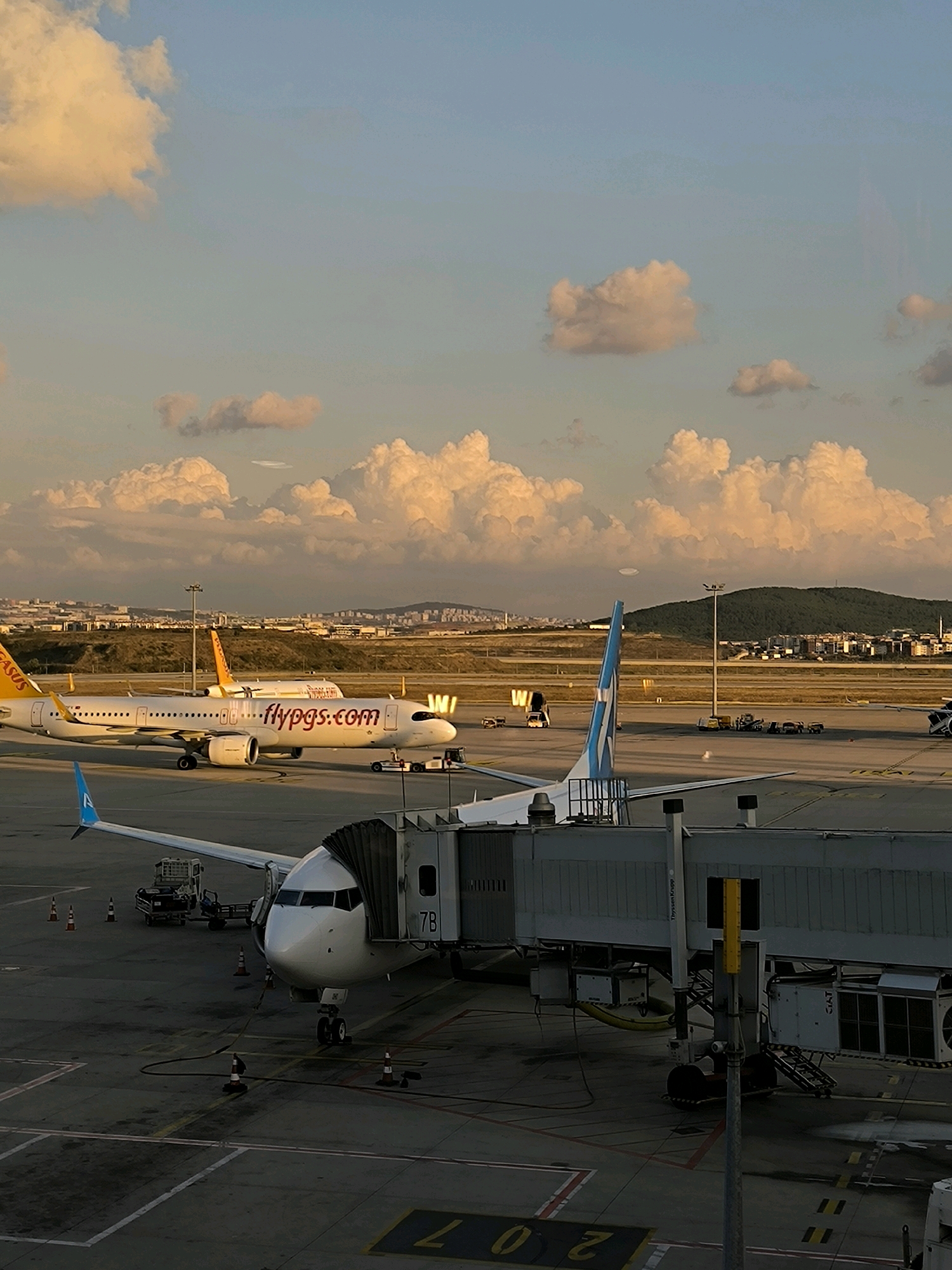 A low-angle, medium shot captures an airport tarmac at dusk in Pendik, Turkey, under a sky filled with dramatic, illuminated clouds. In the foreground, a white passenger jet is connected to a jet bridge. The jet bridge is a long, enclosed walkway, gray in color, with industrial equipment attached. The nose of the aircraft is visible, white with a blue tail fin. Yellow lines mark the tarmac around the aircraft. In the mid-ground, another white passenger jet with "flypgs.com" written in red on its fuselage is parked. This plane has a yellow tail fin and engines. Various ground service vehicles, including baggage carts and tugs, are scattered around the parked aircraft. Beyond the tarmac, a distant city skyline is visible, nestled against rolling, green hills dotted with buildings. The overall lighting suggests the golden hour before sunset.