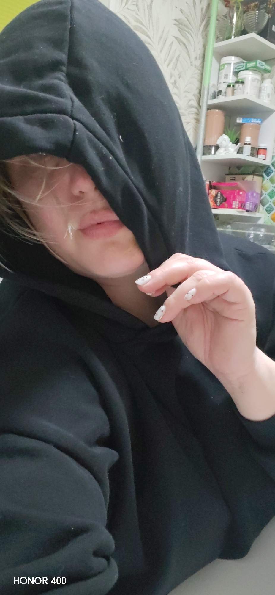 A person wearing a black hooded sweatshirt is shown indoors. The hood obscures part of their face. The person's hand is raised towards their face. A shelf with various items on it is visible in the background. The word "HONOR 400" is visible in the lower-left corner of the image. The city of Noviy, Russia, is not evident in the image.