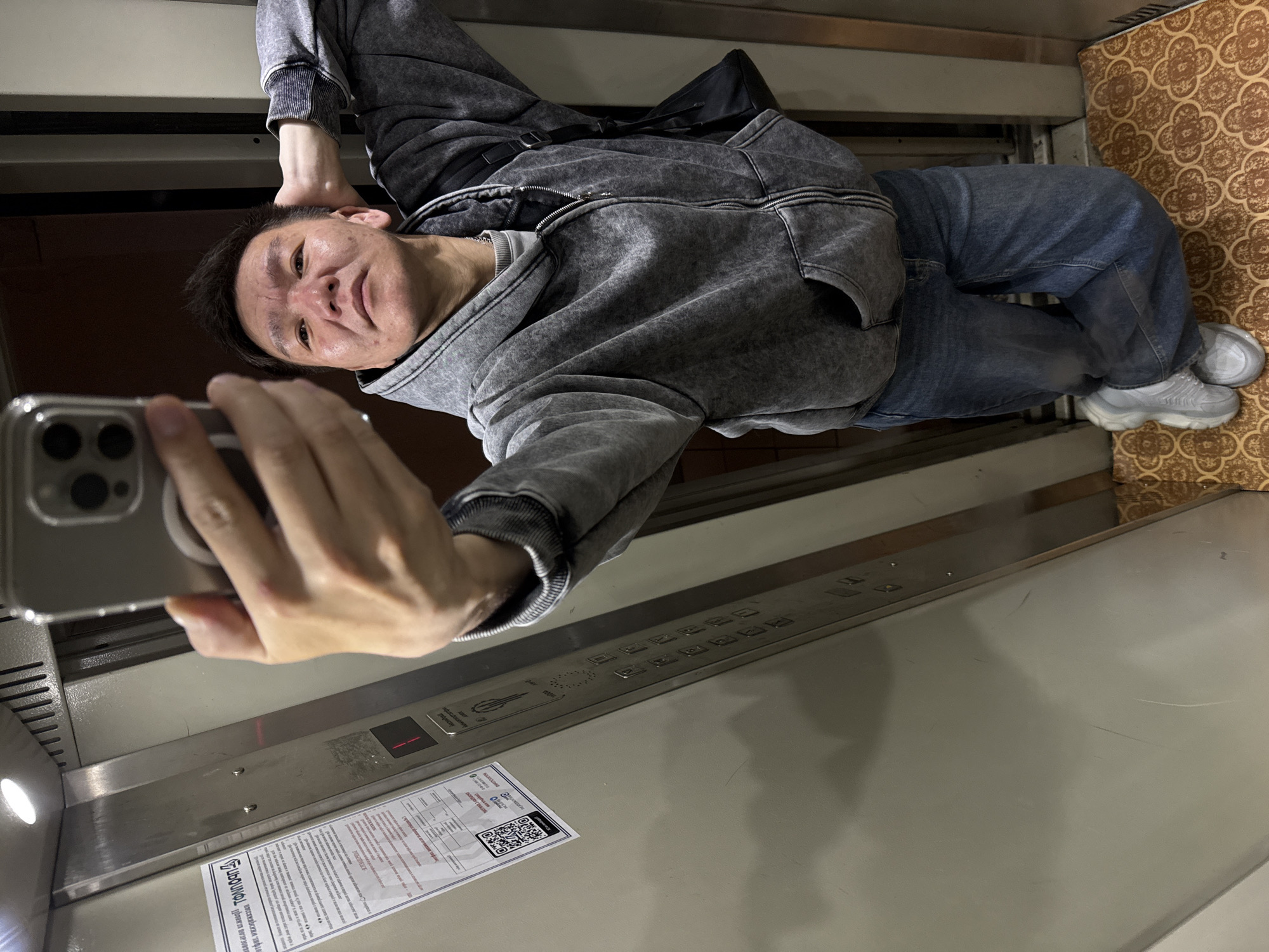 A man stands inside an elevator, taking a selfie with his smartphone. He is wearing a grey hoodie and blue jeans, with grey sneakers visible. A black bag is slung across his body. The elevator has metal walls with a control panel featuring numerous buttons and a QR code. A patterned brown floor is also visible. According to the informational poster inside the elevator (partially visible), the location is in the city of Khimki, Russia.