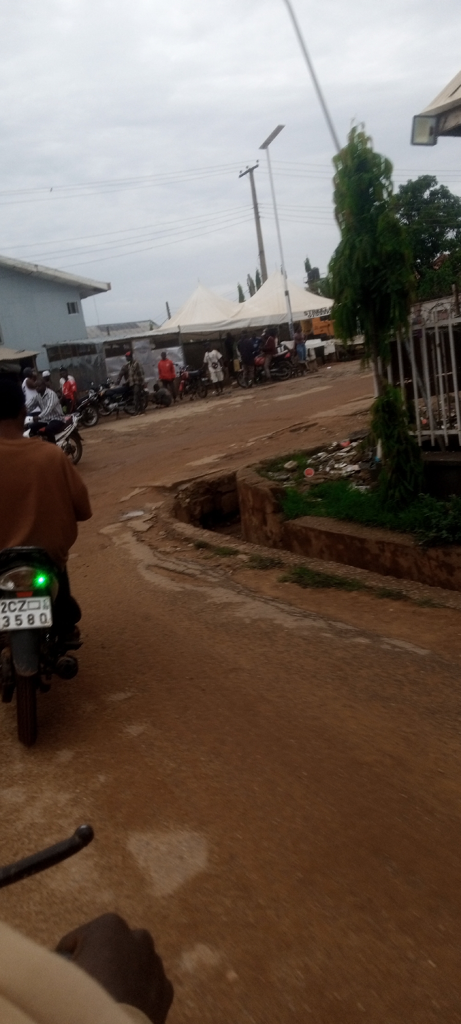 The image, taken from a moving motorcycle in Narayi, Nigeria, shows a dirt road. The motorcycle in front of the camera has a license plate "2CZ 3580". Further down the road, there are people gathered in front of white tents. There are also several parked motorcycles. A building is visible to the left of the tents and a pole is erected above the tents. A tree is to the right of the road. The sky is overcast.