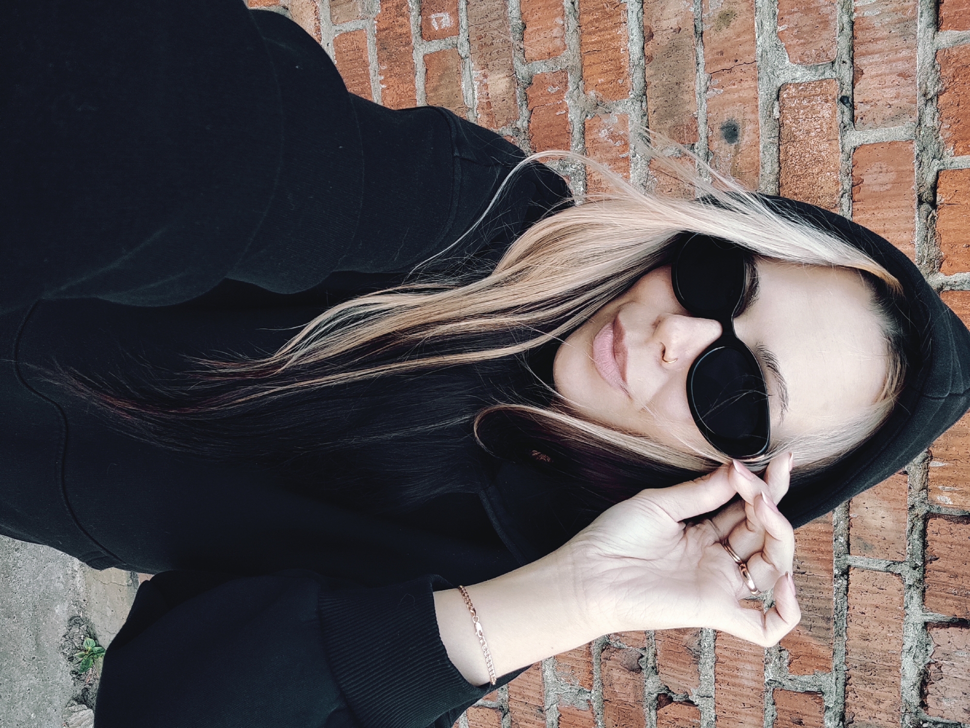 A woman with blonde hair and dark sunglasses is leaning against a brick wall. She is wearing a black hooded sweatshirt. Her left hand is raised near her face, displaying rings and a bracelet. The background is a brick wall with visible mortar. The scene appears to be outdoors, possibly an urban environment. The location is assumed based on provided information and is not directly evident in the image.