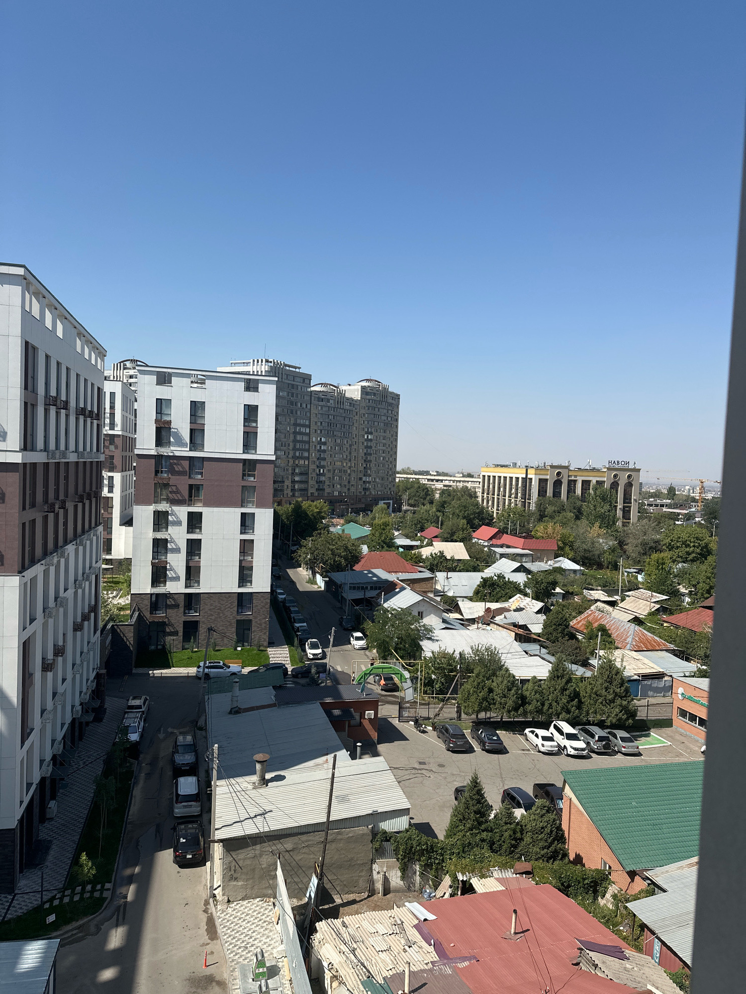 This is a high-angle, vertical, eye-level shot of an urban scene under a clear blue sky. The foreground is dominated by the side of a modern, multi-story apartment building with a white and brown facade, featuring balconies and air conditioning units. To the right of this building, a narrower street runs between it and other residential structures.
Further into the midground, several mid-rise apartment buildings of varying architectural styles are visible, some with many windows and a grayer palette. Interspersed among these buildings are numerous houses with pitched roofs, many appearing to be made of corrugated metal in various shades of red, grey, and brown, suggesting a mix of older and newer construction. Lush green trees are scattered throughout the scene, softening the urban landscape.
In the distance, a large, light-colored building with a distinctive facade and what appears to be signage is prominent, along with other taller buildings that fade into the haze. A construction crane is also visible in the far distance. The street level shows parked cars of various models and colors, and some pedestrian walkways. The overall impression is a dense urban environment with a clear demarcation between modern apartment complexes and more traditional housing.