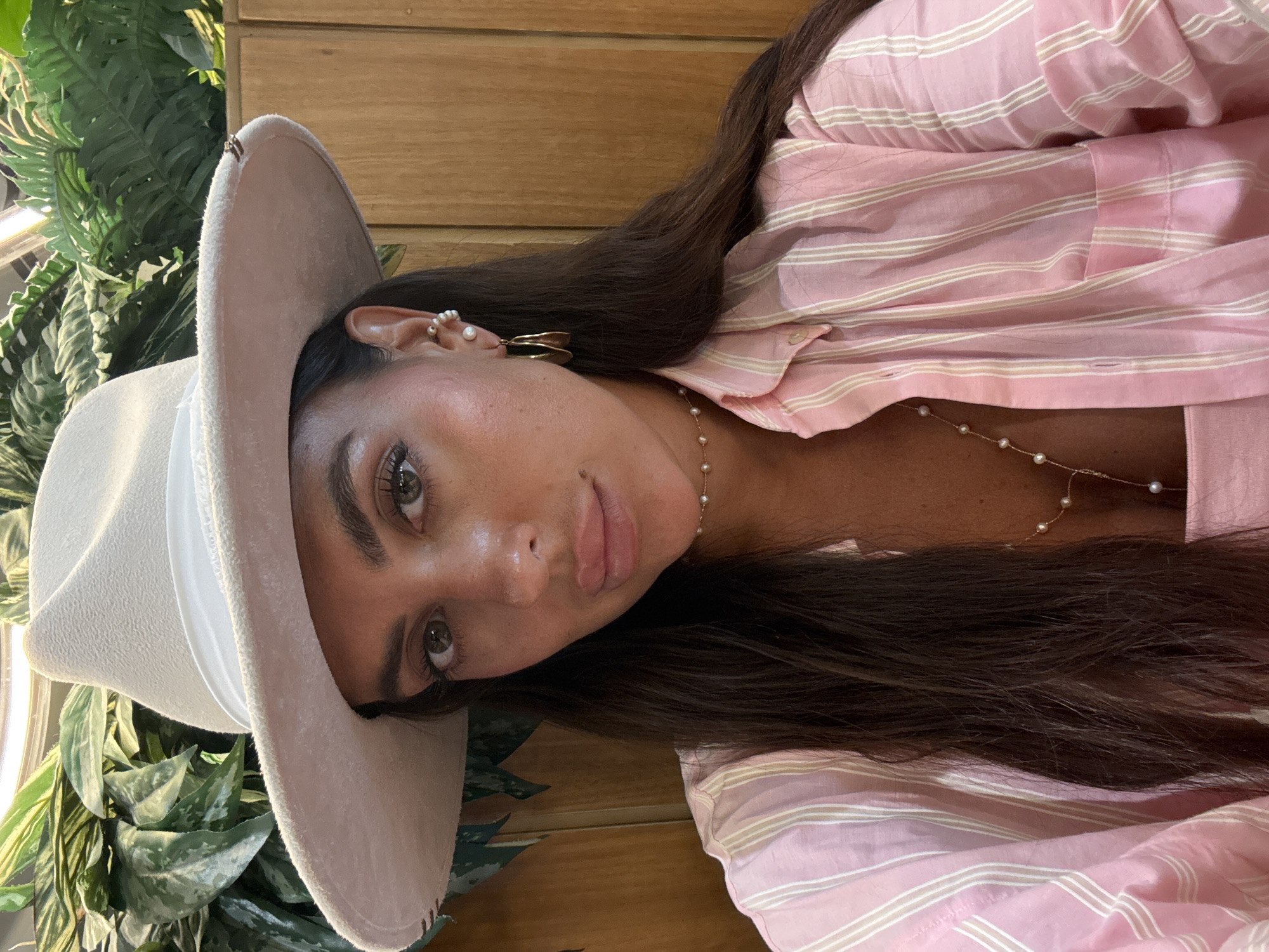 A medium shot captures a woman from the chest up, posing for a selfie. She is wearing a light-colored fedora hat, a pink and white striped button-down shirt, and a delicate pearl necklace. The background consists of lush green foliage and wooden paneling. The lighting suggests an indoor setting, possibly a restaurant or cafe. The woman's makeup is prominent, with defined eyebrows, eyeliner, and lipstick. Her hair is dark brown and falls over her shoulders. She is wearing hoop earrings and another pearl earring on her left ear. The overall aesthetic is stylish and casual. There is no indication of the specific location being in Dubai, United Arab Emirates.