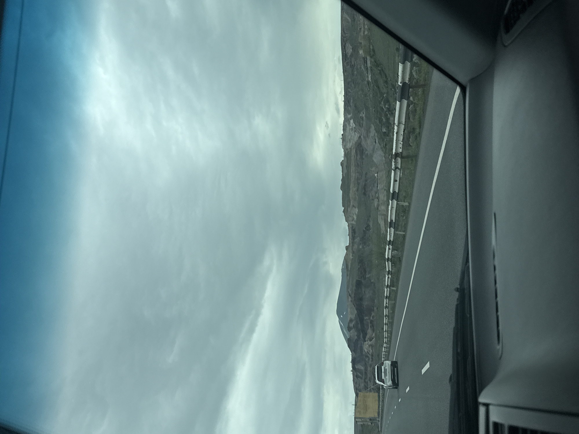 The image depicts a view from inside a vehicle, likely a car, traveling on a highway. The road surface is asphalt, marked with white lines. Adjacent to the highway is a roadside barrier, beyond which is a mountainous landscape. The sky is overcast with a layer of clouds. In the distance, a mountain peak is partially visible. There is a car traveling in the same direction on the road.
Without further evidence, it is impossible to conclusively confirm the exact location as Arzni, Armenia, though the mountainous terrain is consistent with the geography of Armenia.