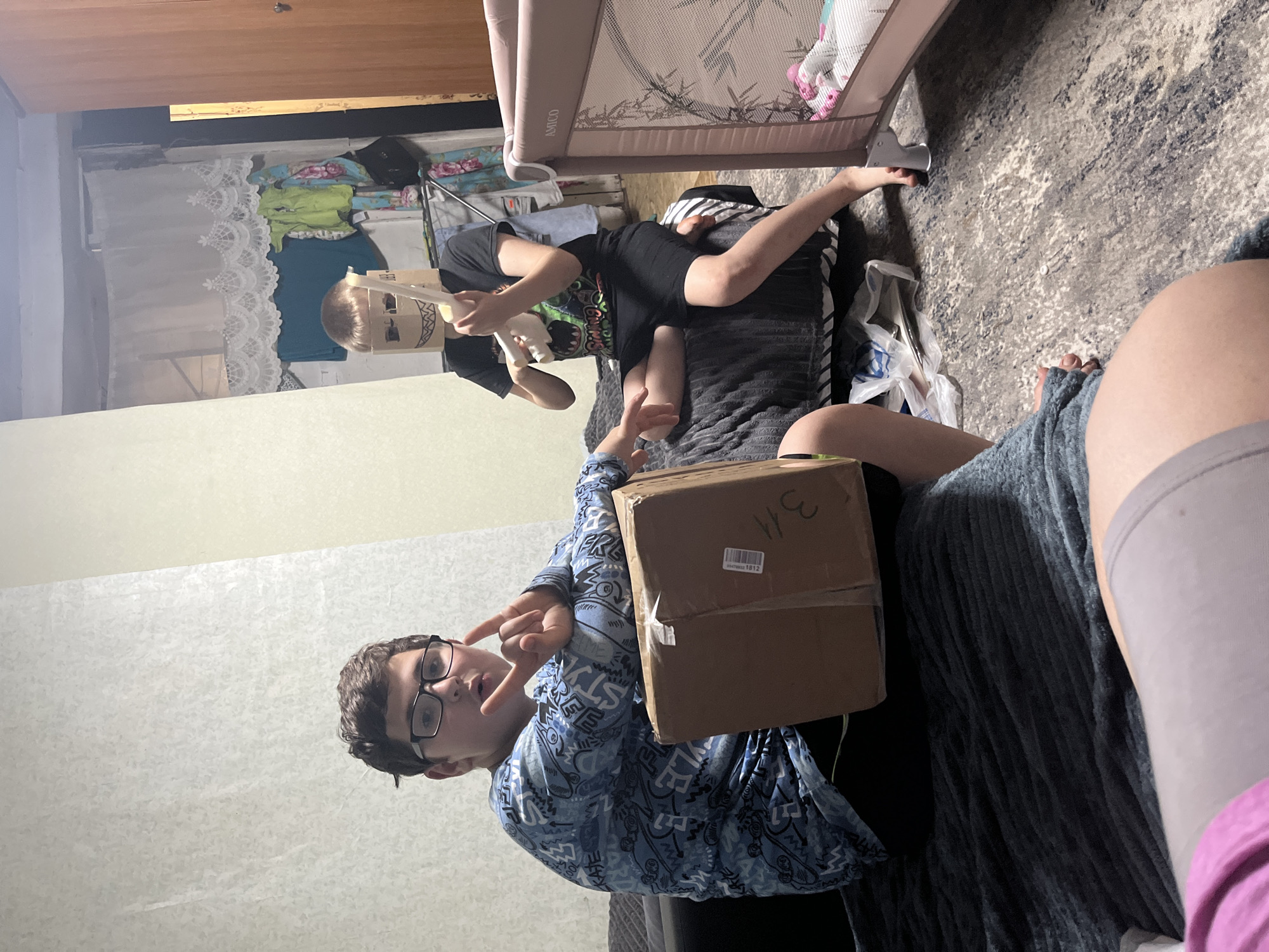 The image captures an indoor residential scene in Фоки, Russia, featuring two primary male subjects and a partial view of a third individual.
On the left, a male with short, dark brown hair and black-framed glasses is seated on a dark gray ribbed blanket covering a sofa or bed. He wears a long-sleeved blue shirt with a repeating abstract or graffiti-like pattern and dark trousers. His right hand is raised, forming a "devil horns" or "rock on" gesture, and his mouth is slightly open. A rectangular brown cardboard box, secured with white tape and marked with "3/4" and an illegible barcode sticker, rests on his lap.
To the right, a second male, appearing younger and with light brown hair, sits cross-legged on the same dark gray blanket. His head is obscured by a makeshift mask fashioned from a light brown cardboard box, featuring cut-out eyeholes and a drawn-on jagged, sharp-toothed mouth. He wears a black t-shirt with a partially visible green graphic containing text, and black shorts, with bare feet. He holds a white, elongated, bone-like or toy weapon object with both hands, positioned in front of him.
The background features light-colored, possibly textured wallpaper. In the mid-ground right, a light pink portable playpen or crib, branded "AMICO," is visible, containing soft bedding and a small pink toy. Behind the second individual, a cluttered area includes a clothes rack with various garments (a white lace valance-like item, a blue shirt, a lime green top, and a floral patterned garment) and a dark brown wooden doorframe. The floor is covered by a gray and white patterned area rug, on which a small white circular object is discernible. Additional plastic bags and miscellaneous items are scattered on the floor near the playpen.
The lower foreground reveals the out-of-focus legs and lap of a third individual, wearing light gray shorts or leggings and a portion of a pink garment. The scene is illuminated by ambient indoor lighting.