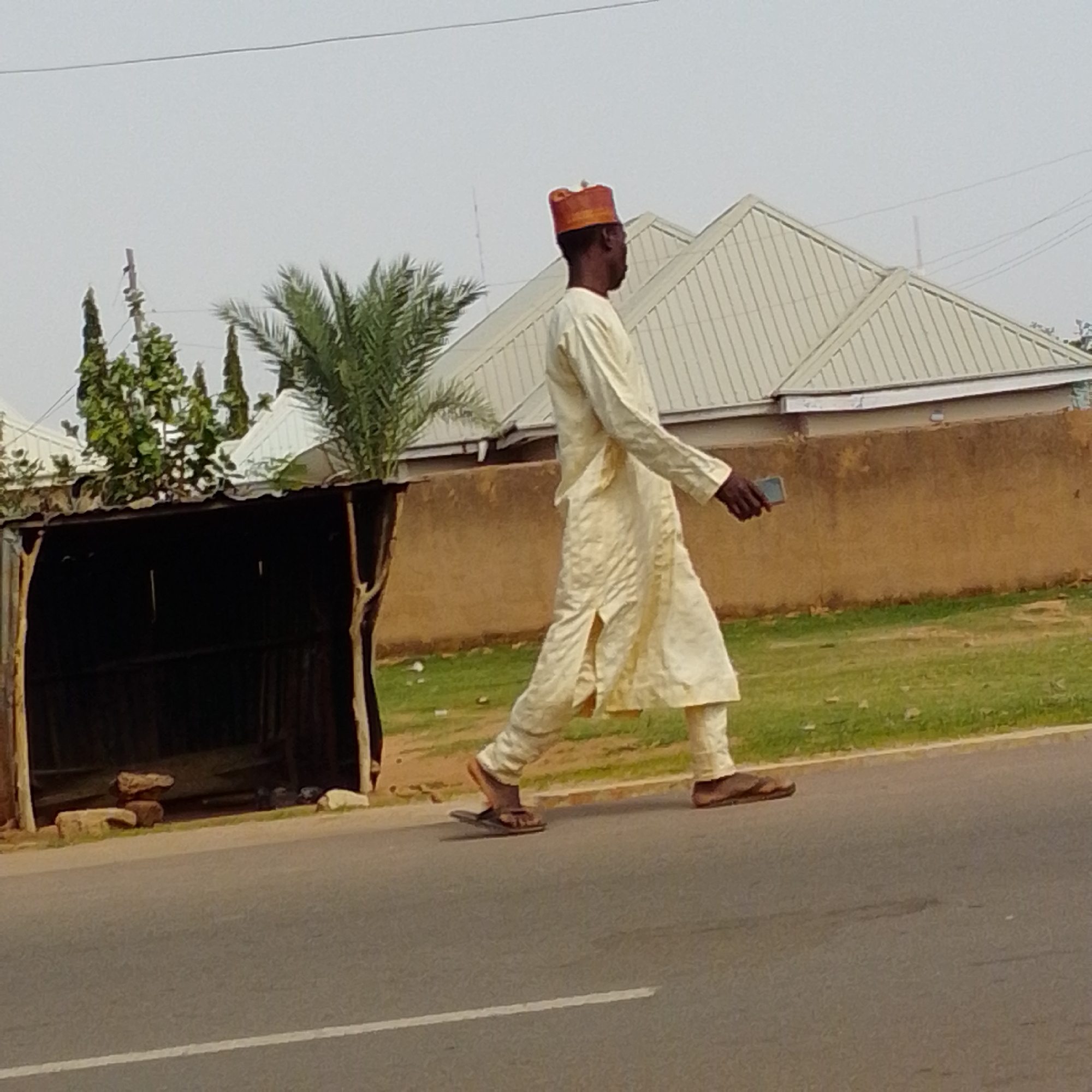 A man wearing a light-colored traditional outfit and an orange cap is walking on a paved road in Gombe, Nigeria. He is holding a dark-colored mobile phone in his right hand. To his left, there is a dilapidated wooden structure with a corrugated metal roof, partially obscured by foliage. Behind the man, a light-colored house with a sloped, ribbed roof is visible, along with some trees and power lines against a hazy sky. The road surface is asphalt with a white line marking. The ground beside the road is covered in grass and dirt.