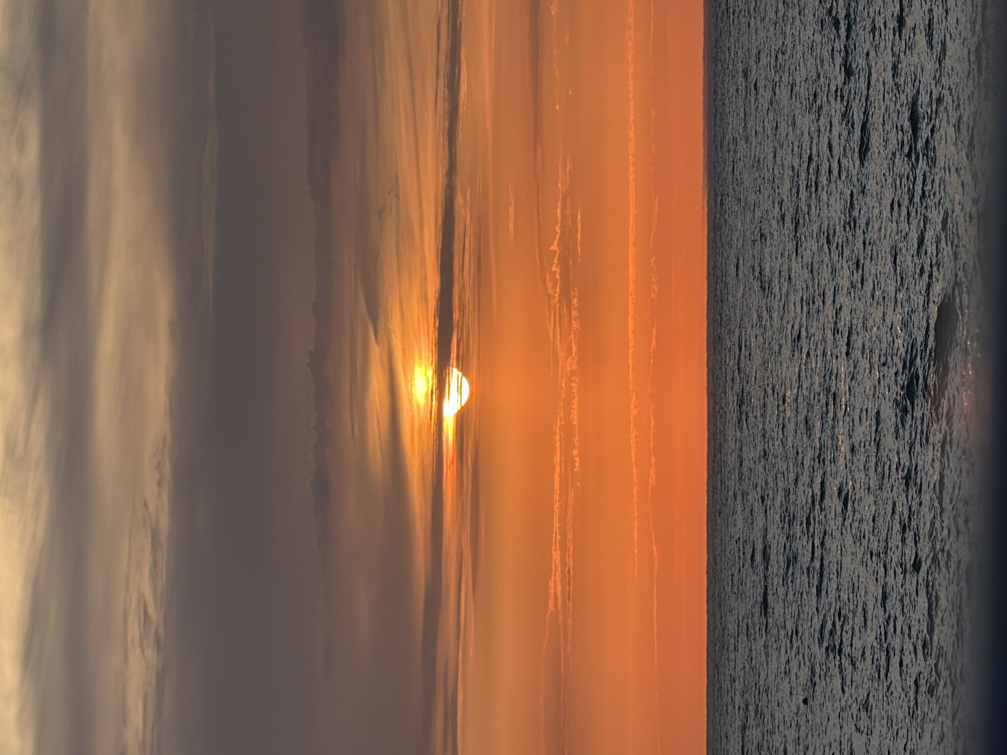 A sunset over the Black Sea is captured from a beach in Sochi, Russia. The sun, partially obscured by clouds, casts a warm orange glow across the sky, with darker clouds gathering at the top. The water is dark and choppy, with gentle waves rolling towards the shore. The horizon is a clear line between the sea and the sky. No people or specific objects are visible in the scene, only the natural elements of the seascape.