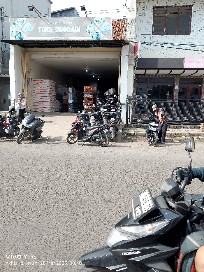 The image shows a street scene in Jatibarang, Indonesia (latitude: -6.475, longitude: 108.307). A row of parked motorcycles are parked in front of a store with a sign that says "TOKO SODADI" and "TOKO PLASTIK & BAHAN RUI". There is a man standing in the middle of the image, looking down, presumably adjusting something on one of the parked motorcycles. There are two more people standing in the doorway of the store, but their faces are not visible. The image is captured from a low angle, facing the front of a black Honda motorcycle parked in the foreground. The motorcycle is facing away from the camera. The street is paved with asphalt and appears to be in good condition. There are no visible cars in the image. The building behind the parked motorcycles has a white brick facade and a brown awning above the doorway. The image is shot in a bright, sunny, daytime setting.