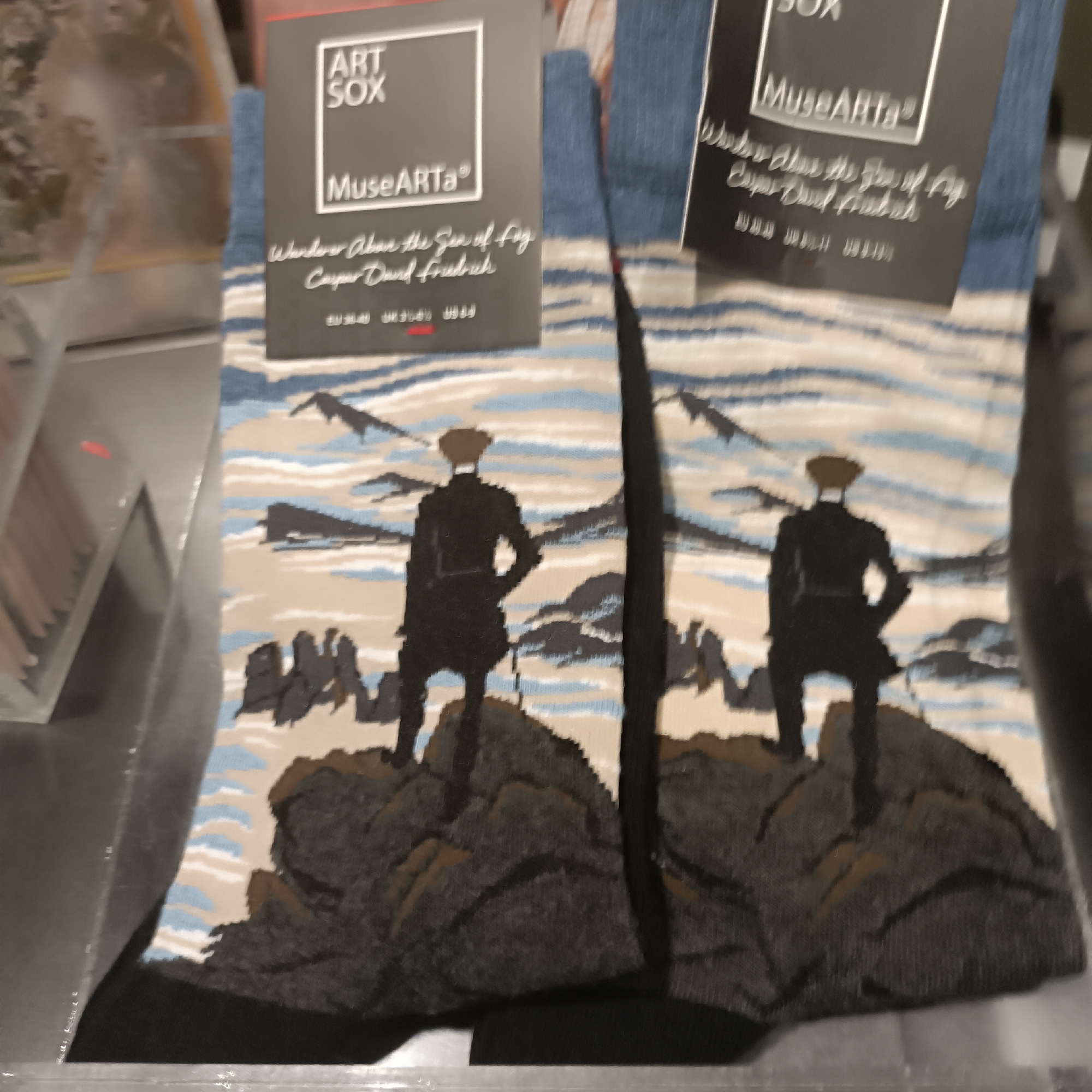 The image displays a pair of socks with a printed design depicting two figures in silhouette standing on rocky outcrops, facing a misty, mountainous landscape. The socks are folded and presented as a product, with packaging tags visible. One tag reads "ART SOX" and "MuseARTA®", with the text "Wanderer Above the Sea of Fog" and "Caspar David Friedrich" printed below. The other tag has the "ART SOX" logo. The socks themselves feature a pattern of wavy lines in blues, whites, and tans, interspersed with darker mountain shapes and the silhouettes of the figures. The setting appears to be a retail environment, possibly a store display, given the presentation of the product and the blurred background suggestive of shelves or merchandise. There is no indication of the specific location within the city of Frankfurt, Germany.
