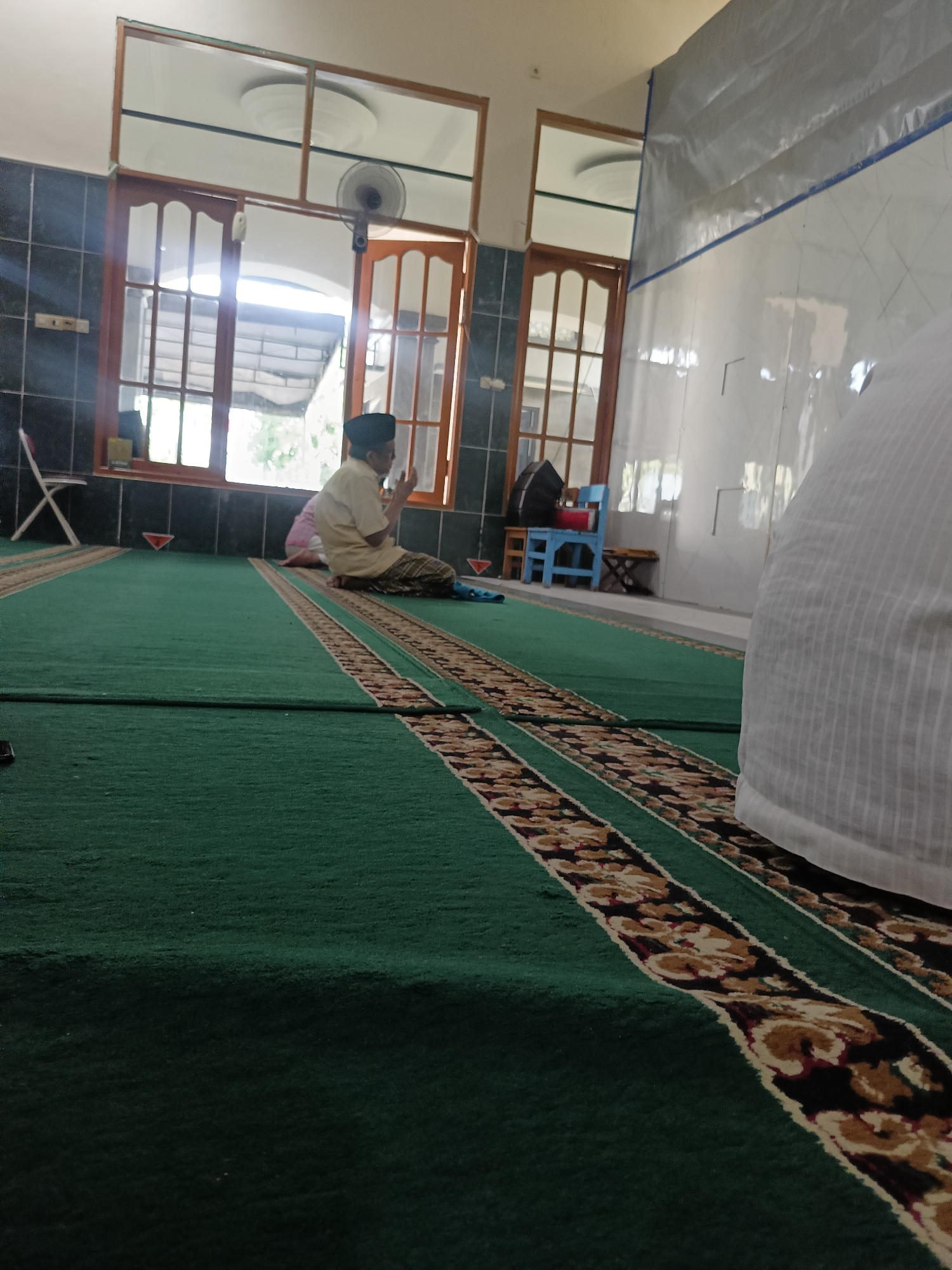 A male, wearing a yellow shirt, black cap, and sarong, is praying on a green carpet with a patterned border inside a room. He is sitting with his hands raised. Sunlight streams through large wooden-framed windows, illuminating the room. The walls are tiled in black and white. In the background, a white object, possibly a prayer mat or a piece of fabric, is partially visible. A blue chair and a small wooden stool are also present. The scene appears to be in a mosque or prayer room in Kediri City, Indonesia.