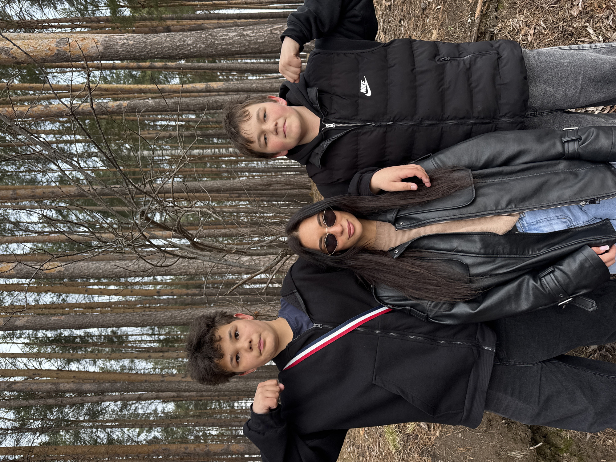Three individuals are positioned in front of a dense pine forest. The person at the top is wearing a black Nike puffer vest, jeans, and a hooded top. The person in the middle is wearing sunglasses and a black leather jacket. The person at the bottom is wearing a black jacket with a red, white, and blue stripe on the shoulder and black pants. The location is in a forest in the city of Chaikovsky, Russia.