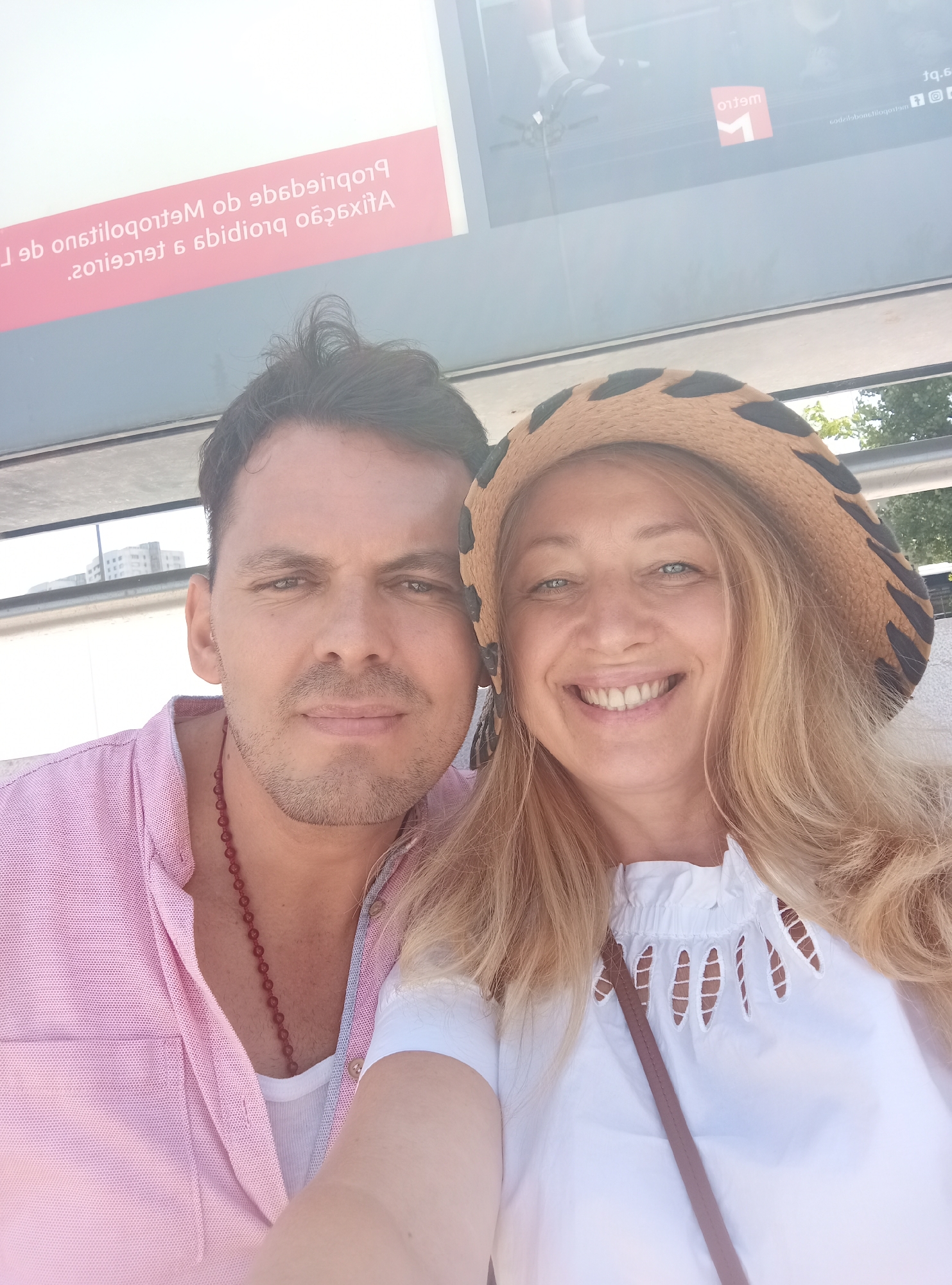 A close-up selfie features a man and a woman posing in what appears to be a public transportation waiting area in Lisbon, Portugal. The man, on the left, is wearing a pink button-down shirt and a red beaded necklace. The woman, on the right, is wearing a wide-brimmed hat with a leopard print pattern and a white ruffled top. They are both smiling and looking at the camera. Behind them, a sign with text in Portuguese is visible, along with the logo of Metro de Lisboa. The environment suggests an outdoor setting, possibly a bus stop or metro station platform, with a building visible in the background.