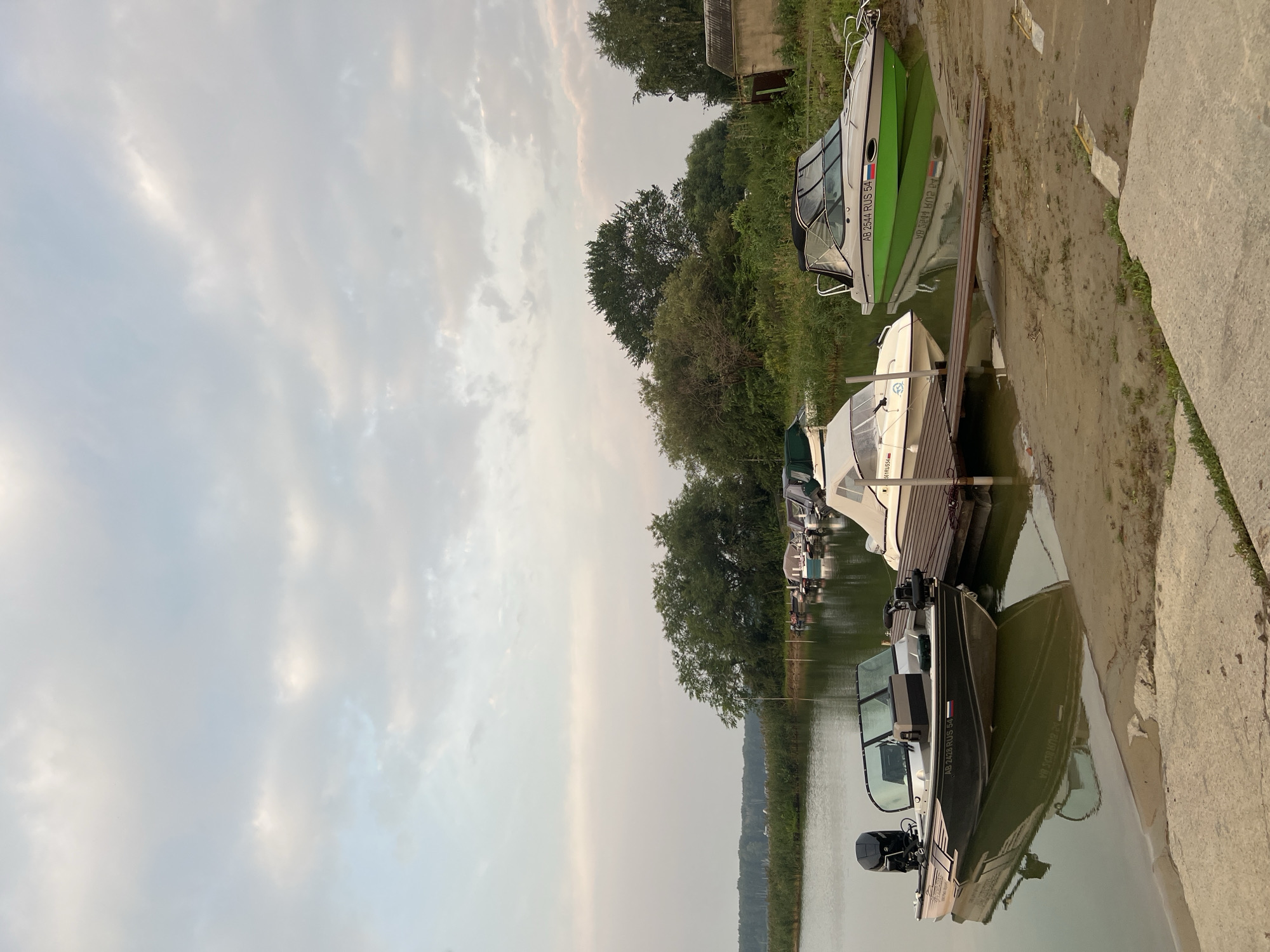 The image depicts a serene waterfront scene in Novopichugovo, Russia. In the foreground, a dark-colored motorboat with an outboard motor is moored to a concrete ramp. To the right, a bright green and white motorboat is docked on a wooden pier. Several other boats, some covered, are visible further down the pier, suggesting a small marina. The background is dominated by lush green trees and reeds lining the water's edge. The sky is overcast with soft clouds, casting a diffused light over the scene. The overall impression is one of a peaceful, perhaps early morning or late evening, setting in a quiet, natural environment.