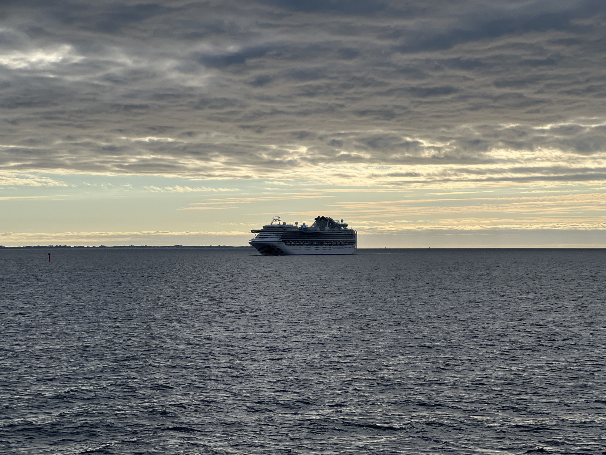 A large cruise ship is seen in the distance on a dark, choppy sea under a heavily clouded sky. The ship is white with blue accents and has multiple decks and structures. In the foreground, the dark water fills the lower half of the frame. On the horizon, a thin strip of land with some buildings is visible, along with a red buoy. The sky is filled with layers of grey and white clouds, with a band of lighter sky near the horizon where the sun appears to be setting or rising. This scene is likely the Bay of Gdynia, Poland, given the presence of a cruise ship and the characteristic coastal landscape.