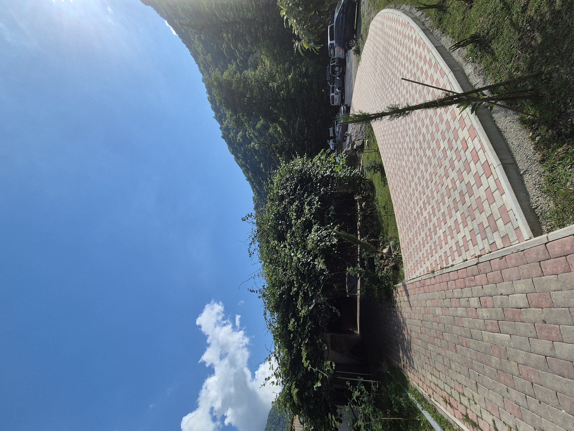 A paved walkway with a red and white checkerboard pattern curves around a grassy area in a mountainous region. Several cars are parked along the edge of the walkway, facing away from the camera, suggesting a parking area. A lush, green mountain slope covered in trees rises steeply to the left of the walkway. The sky is a bright, clear blue with a few wispy white clouds. The sun is shining brightly, casting a slight glare on the upper left portion of the image, indicating it is daytime. The overall scene is one of a scenic outdoor location, likely a tourist spot or a resting area within a natural landscape. The presence of a paved walkway and parked cars suggests some level of development in the area. There is no visible text in the image.