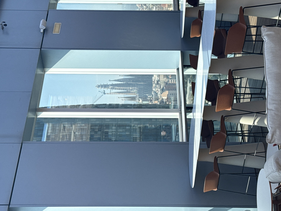 The image depicts an interior space, likely a modern lounge or communal area, located on an upper floor of a building in Barcelona, Spain. The room features blue-grey walls and large floor-to-ceiling windows. In the midground, several long, light-colored high-top tables, supported by white conical bases, are arranged with multiple high-back bar stools that have brown/orange fabric seats and black metal frames. A light-colored sofa section and a small round table are partially visible in the foreground. A white security camera and a rectangular fixture are mounted on the wall above the window frame on the right.
Through the large windows, the iconic Sagrada Familia cathedral is prominently visible in the distance, characterized by its multiple spires and an active construction crane near its top. The surrounding cityscape of Barcelona, consisting of dense residential and commercial buildings, extends below the cathedral. A reflection of a tall, modern building with a grid-patterned facade is visible in the left portion of the window glass. Within this reflective area or through an adjacent internal glass partition, a solitary individual in dark clothing, appearing to be seated at a workstation, is partially visible. No active interactions or specific actions are discernible.