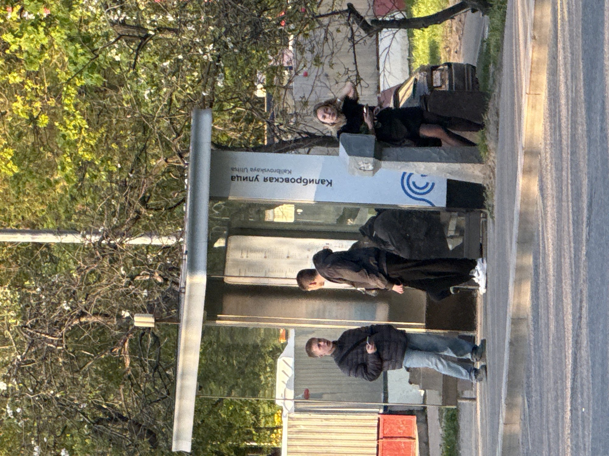 The image depicts a bus stop on Kalibrovskaya Ulitsa in Moscow, Russia. Three individuals are visible at the stop. One person is standing and holding a phone, while another is standing to the left of the bus stop. A third person is lying down on the bus stop bench. The environment shows a sidewalk and trees.