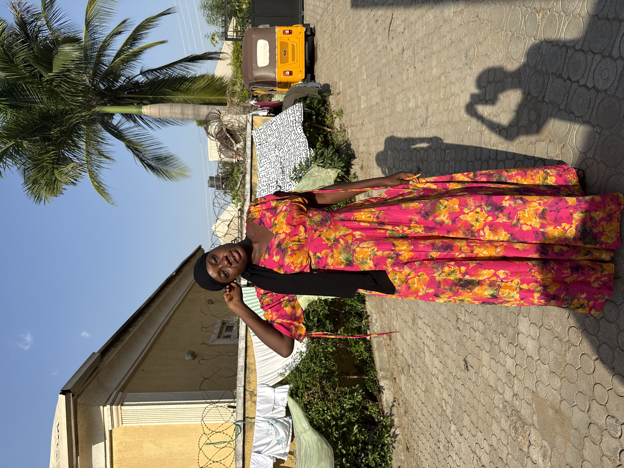 A woman wearing a black head covering and a long, pink and yellow floral dress stands outside on a patterned stone surface. A shadow suggests someone is taking her photo. Behind her are a yellow building with barbed wire, a palm tree, a yellow auto rickshaw, and hanging laundry. The location is inferred to be Abuja, Nigeria.